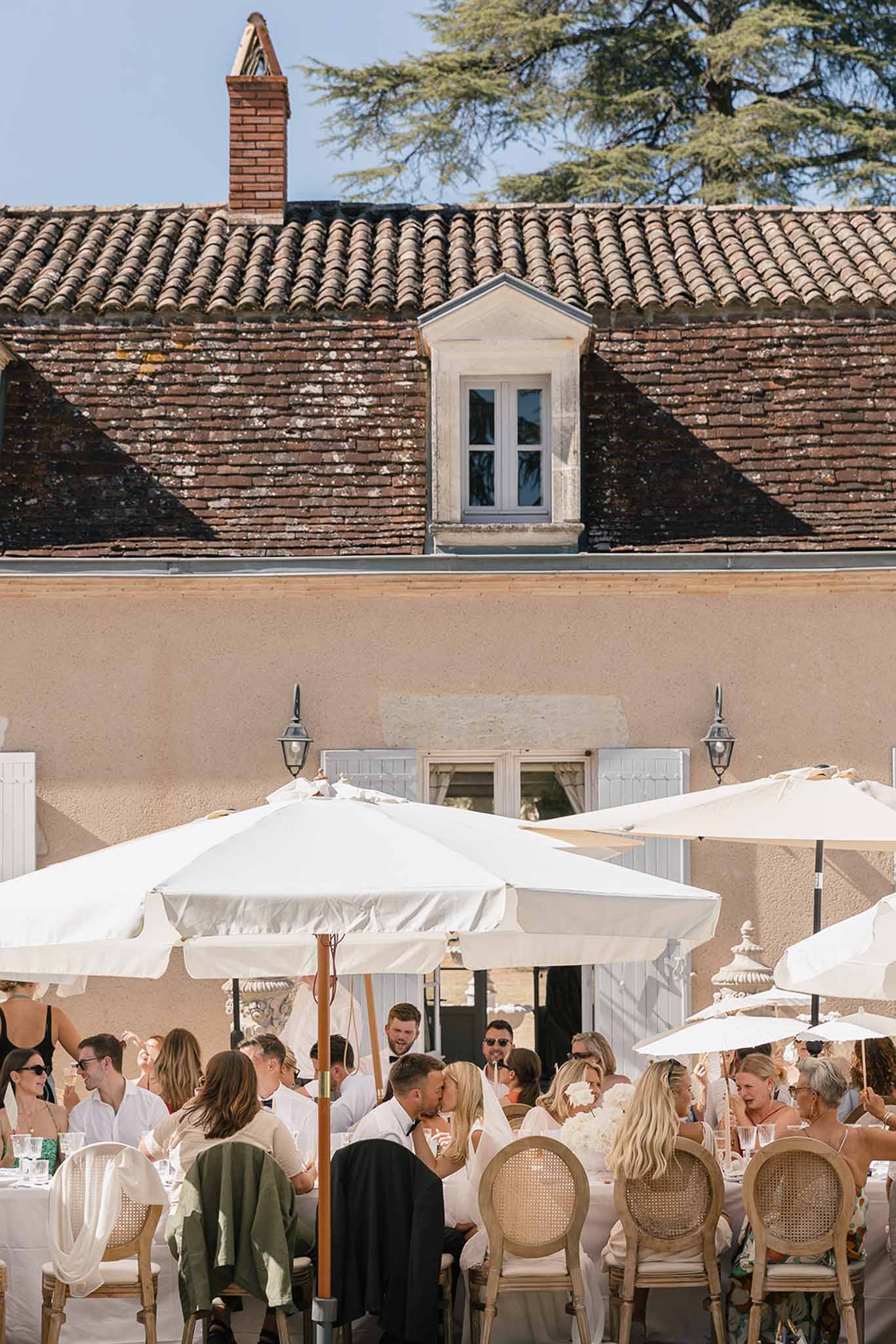 Couple kissing among 25 guests at long table with white umbrellas before terracotta-roofed manor