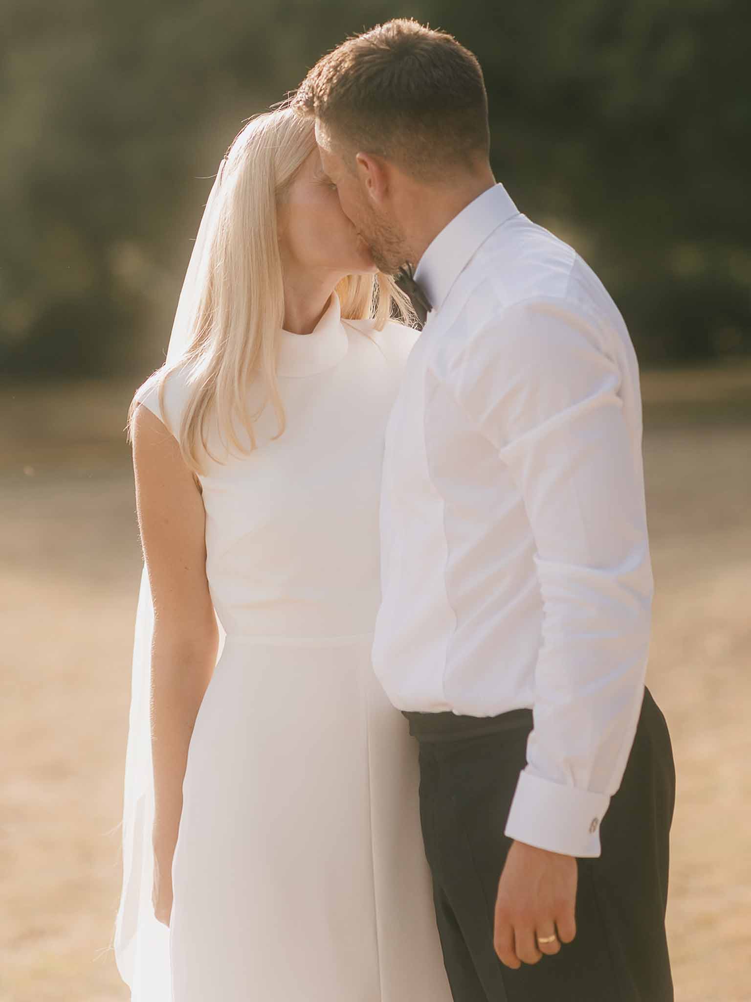 Bride in ivory cap-sleeve gown and cathedral veil kissing groom in golden sunset light outdoors