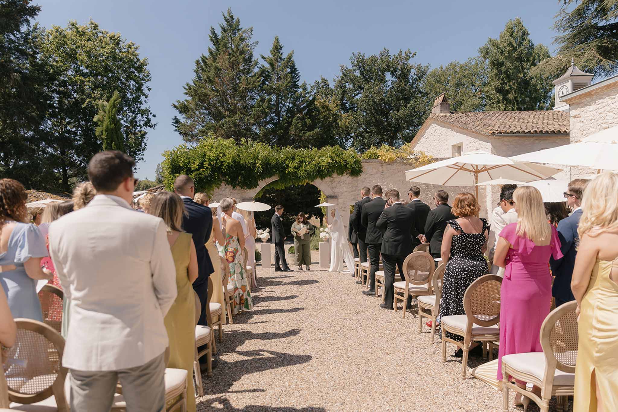 Outdoor wedding ceremony on gravel courtyard with guests seated under cream parasols facing bride and groom