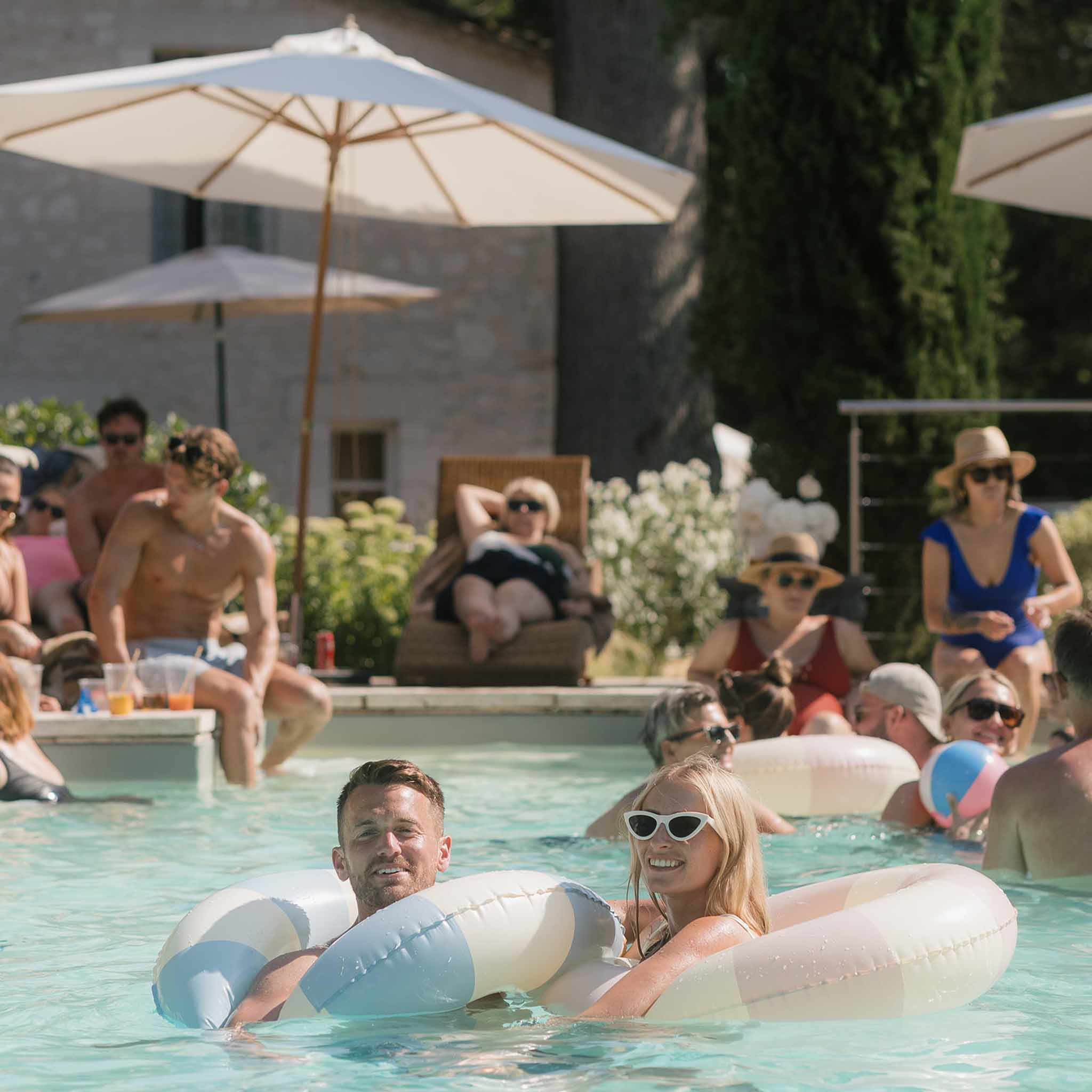 Guests swimming and lounging on pool floats beside stone farmhouse with parasols at wedding after-party