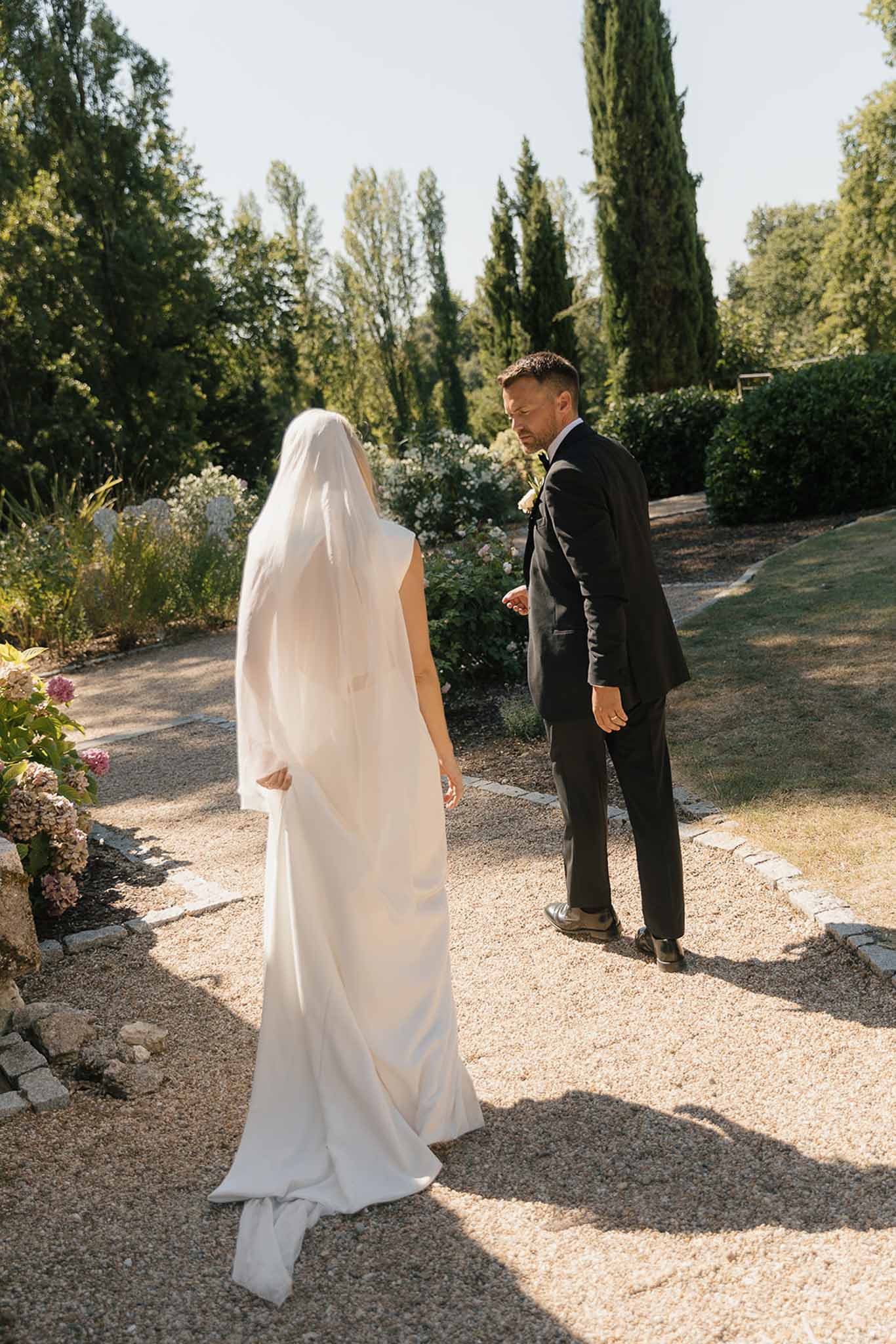 First look on garden path as veiled bride approaches groom among cypress trees and hydrangeas