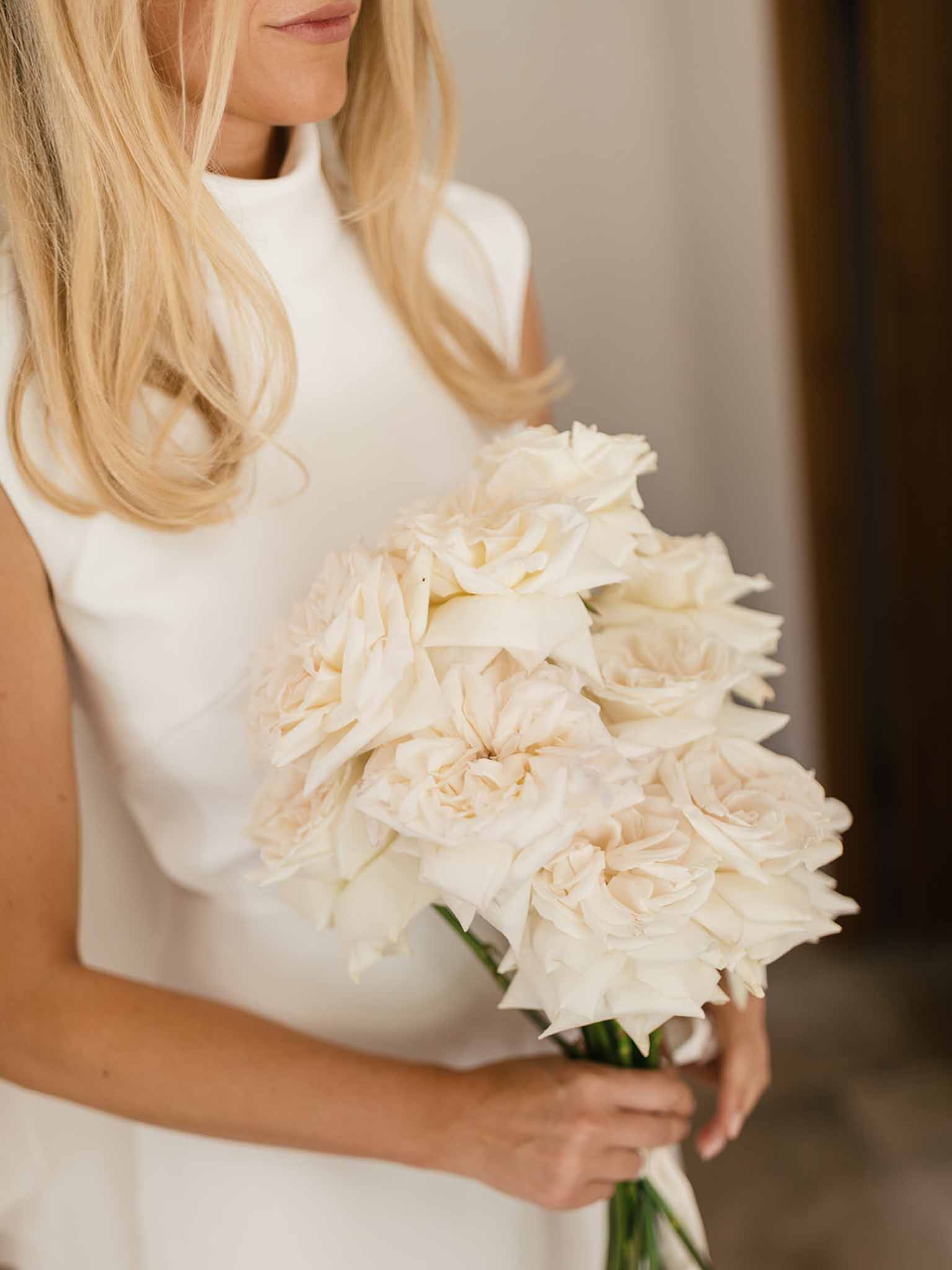 Bride in white mock-neck sleeveless gown holding tight round bouquet of cream and blush garden roses indoors