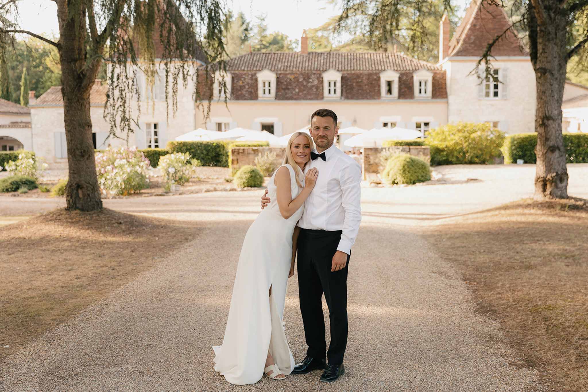 Bride in ivory crepe gown leaning into groom in tuxedo on chateau gravel drive at golden hour