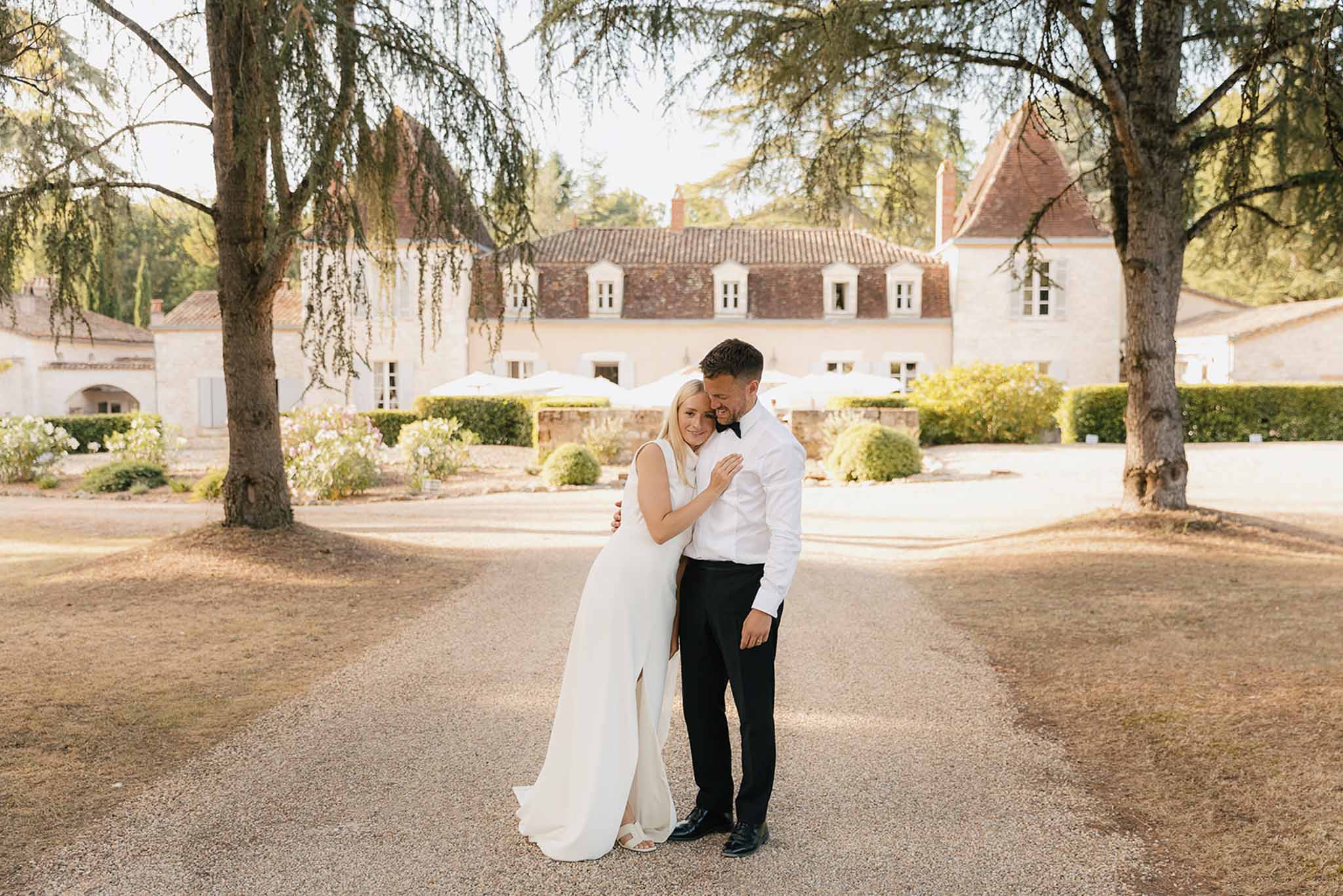 Couple embracing on gravel driveway at golden hour bride in minimalist white gown groom in black tuxedo
