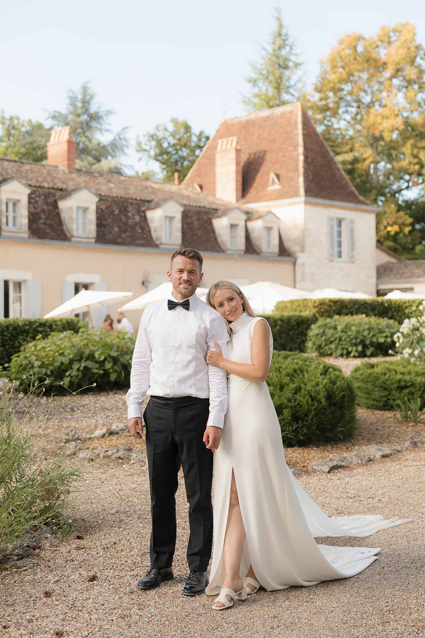 Bride in sleeveless ivory crepe gown leaning on groom in black bow tie on gravel path outside French chateau