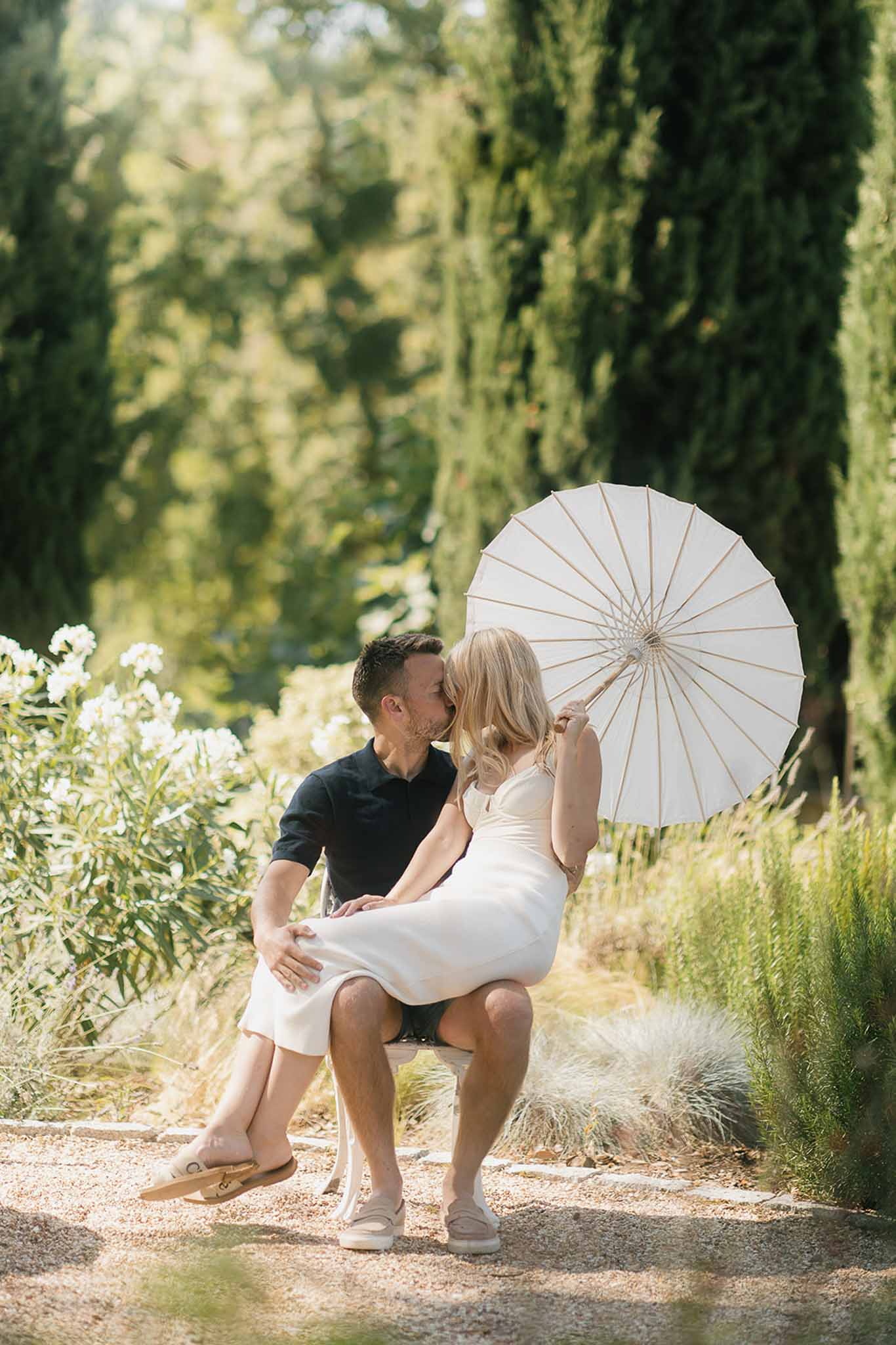 Couple kissing on garden bench along cypress-lined gravel path, woman in ivory slip dress holding white parasol