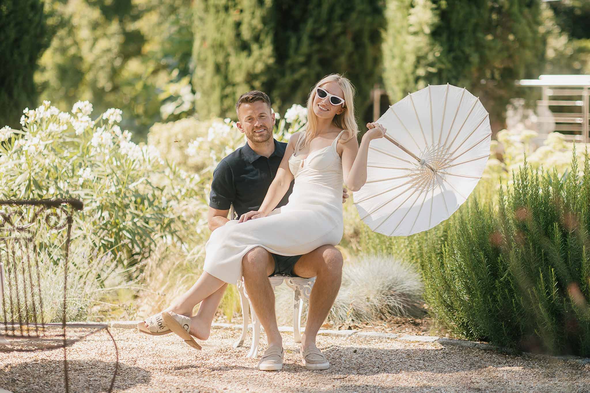 Relaxed couple with bride in slip dress and sunglasses sitting on groom's lap holding white parasol
