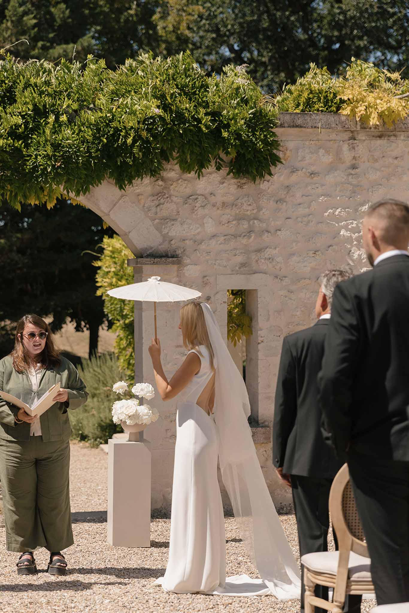 Outdoor courtyard wedding ceremony with bride in white jumpsuit and cathedral veil holding parasol at limestone archway altar