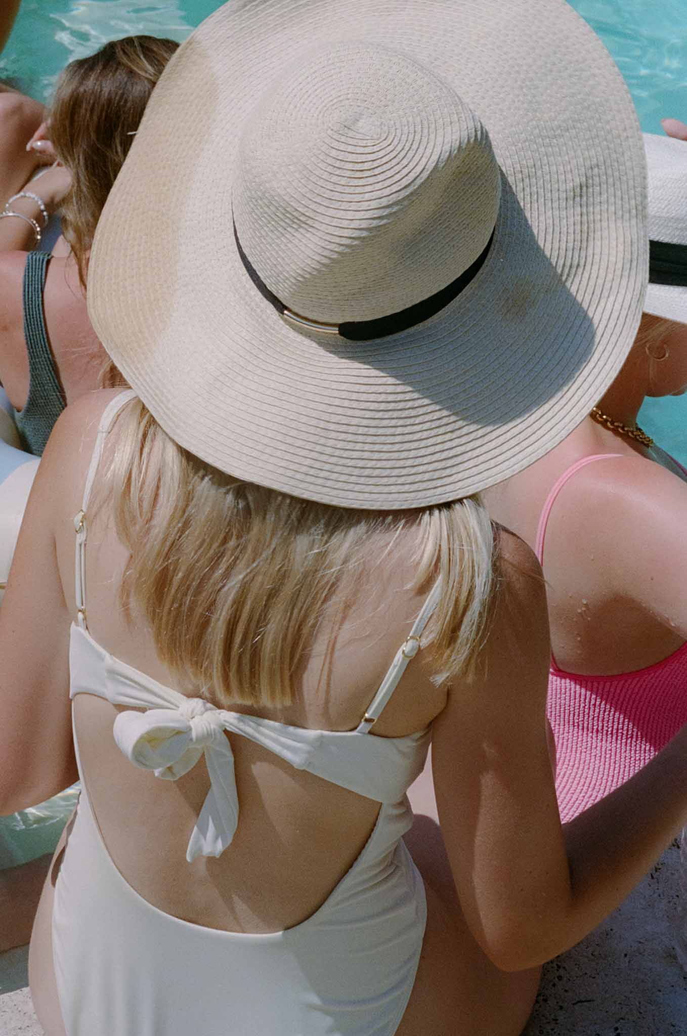 Bride and two friends seated at poolside edge wearing swimsuits and sun hats at pre-wedding gathering