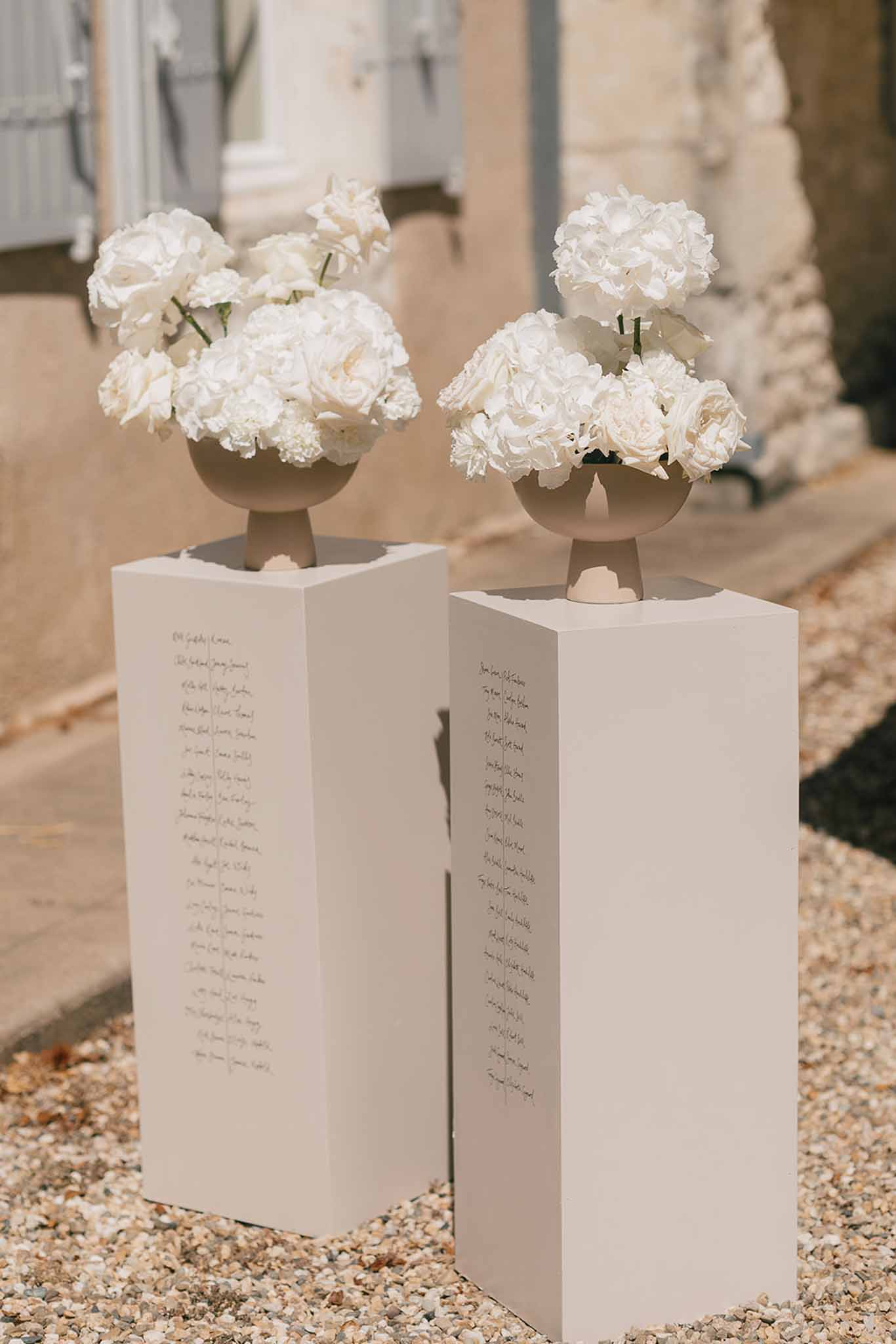 White pedestal seating chart displays topped with white rose and hydrangea arrangements outside French stone building