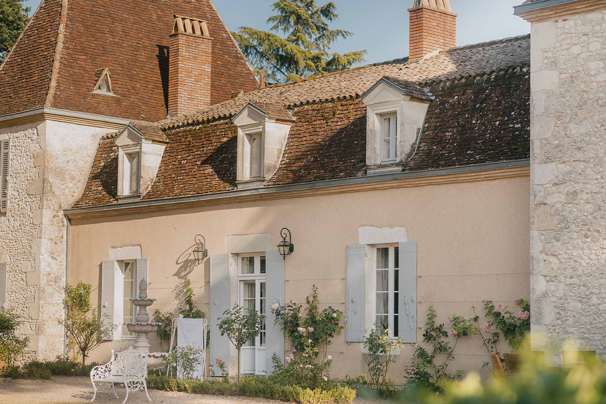 French manor with peach facade, blue shutters, climbing pink roses, and tiered stone fountain
