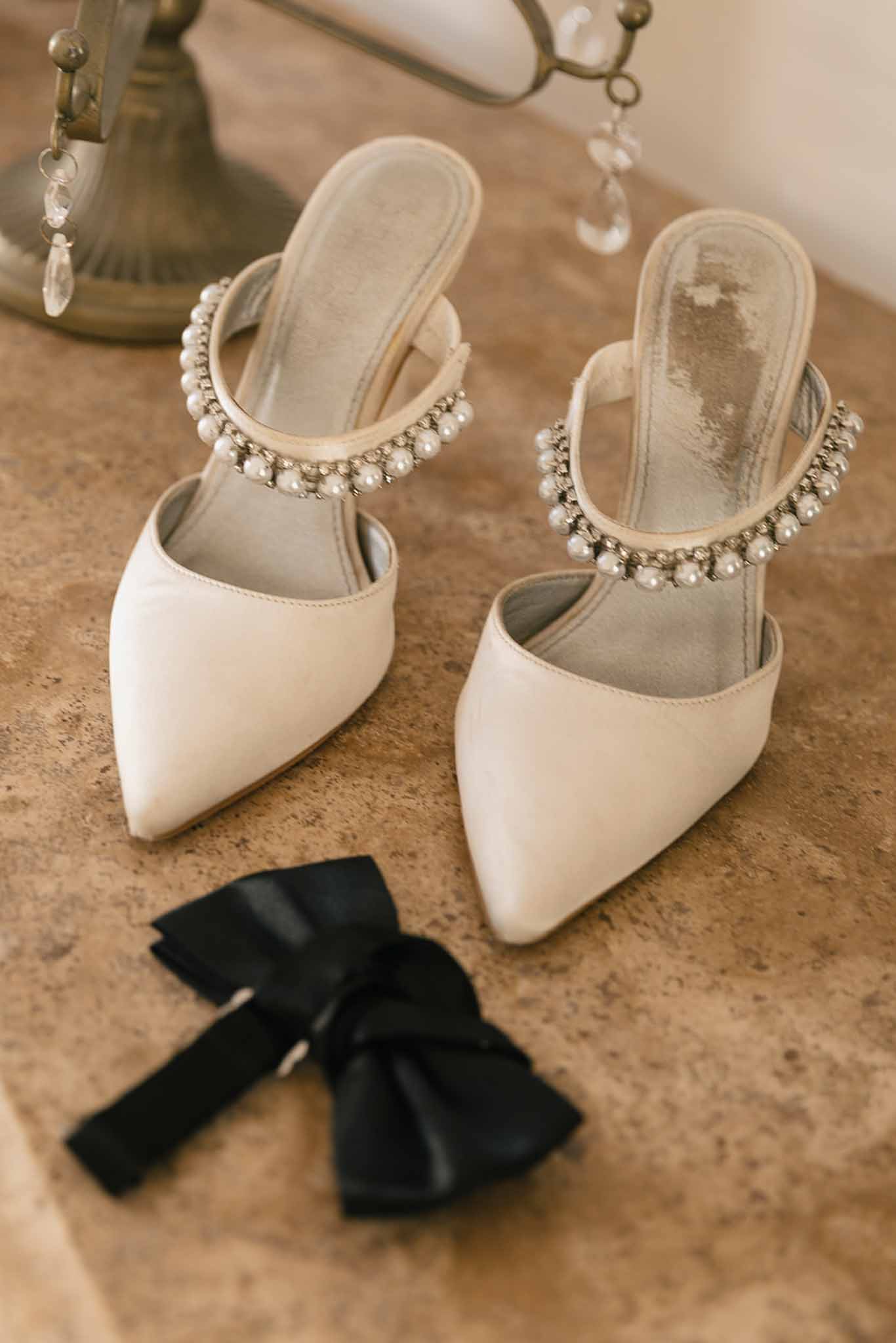 Ivory satin pointed-toe mule heels with pearl ankle straps and black satin bow on stone surface