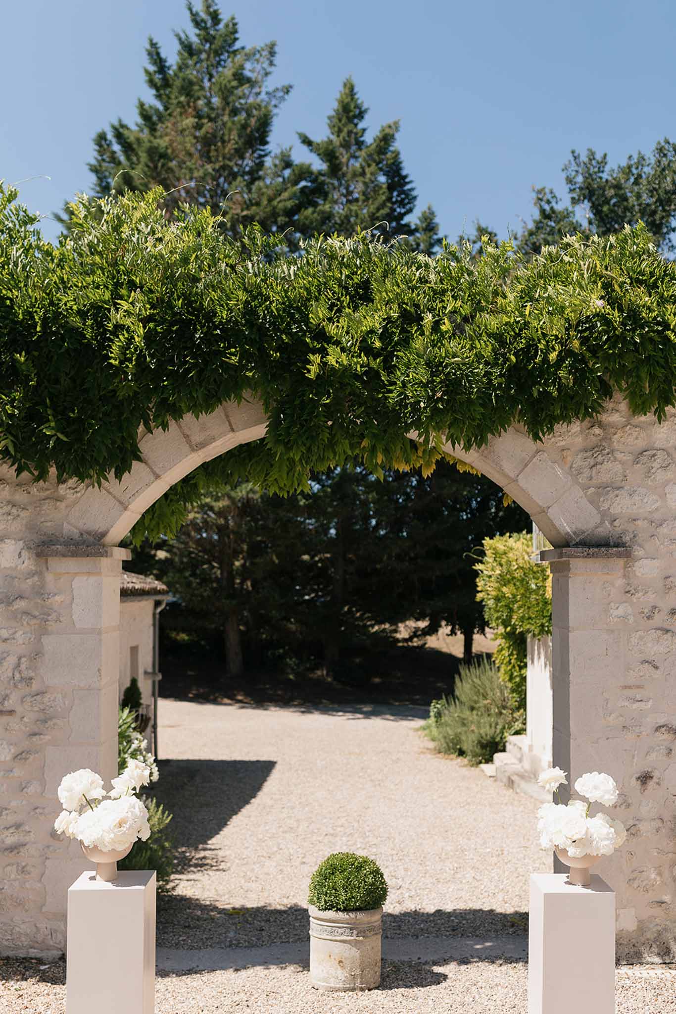 Stone archway entrance with white peony arrangements in urns and boxwood topiary on white pedestals