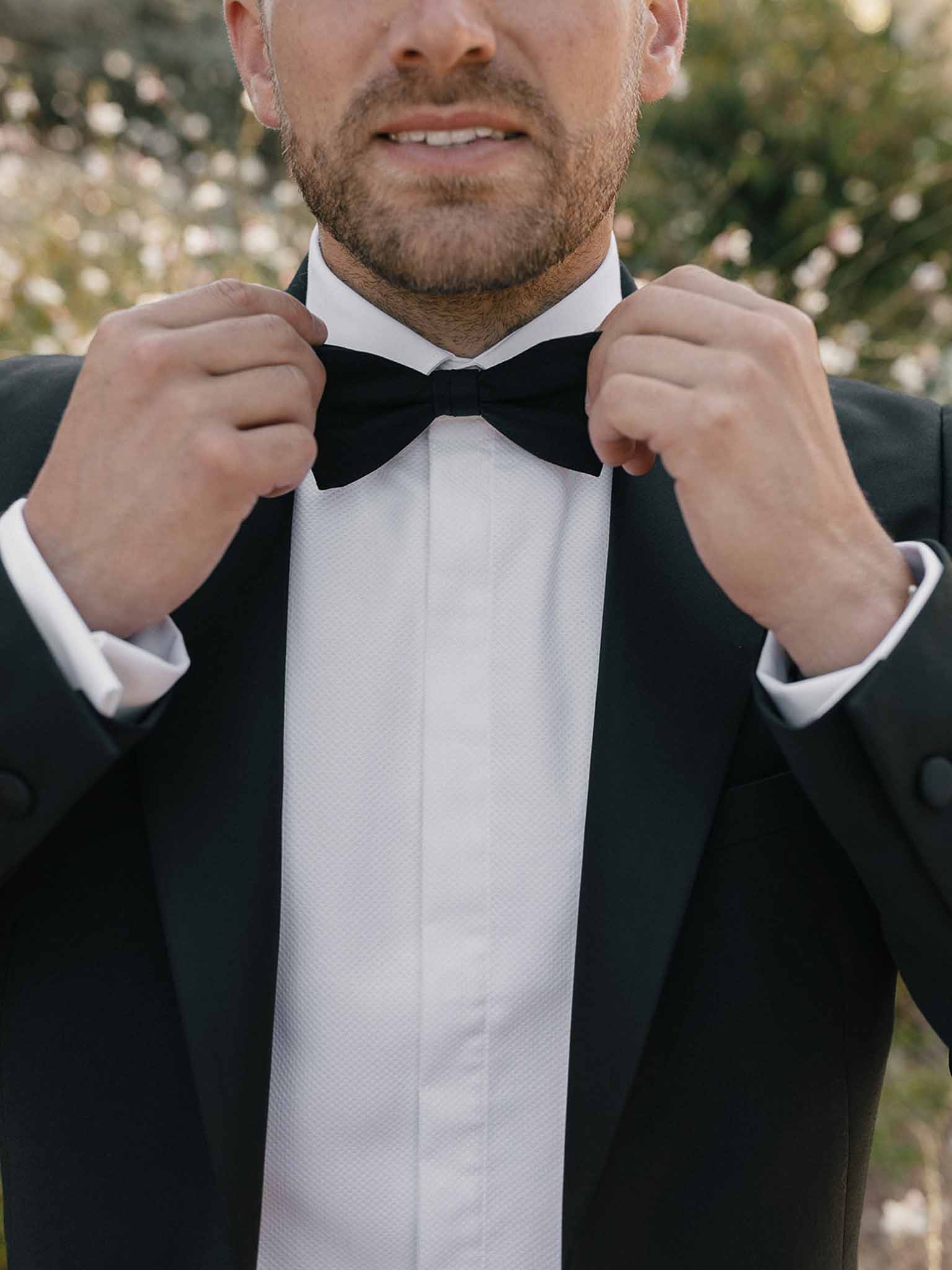 Groom adjusting black velvet bow tie wearing classic tuxedo with satin lapels