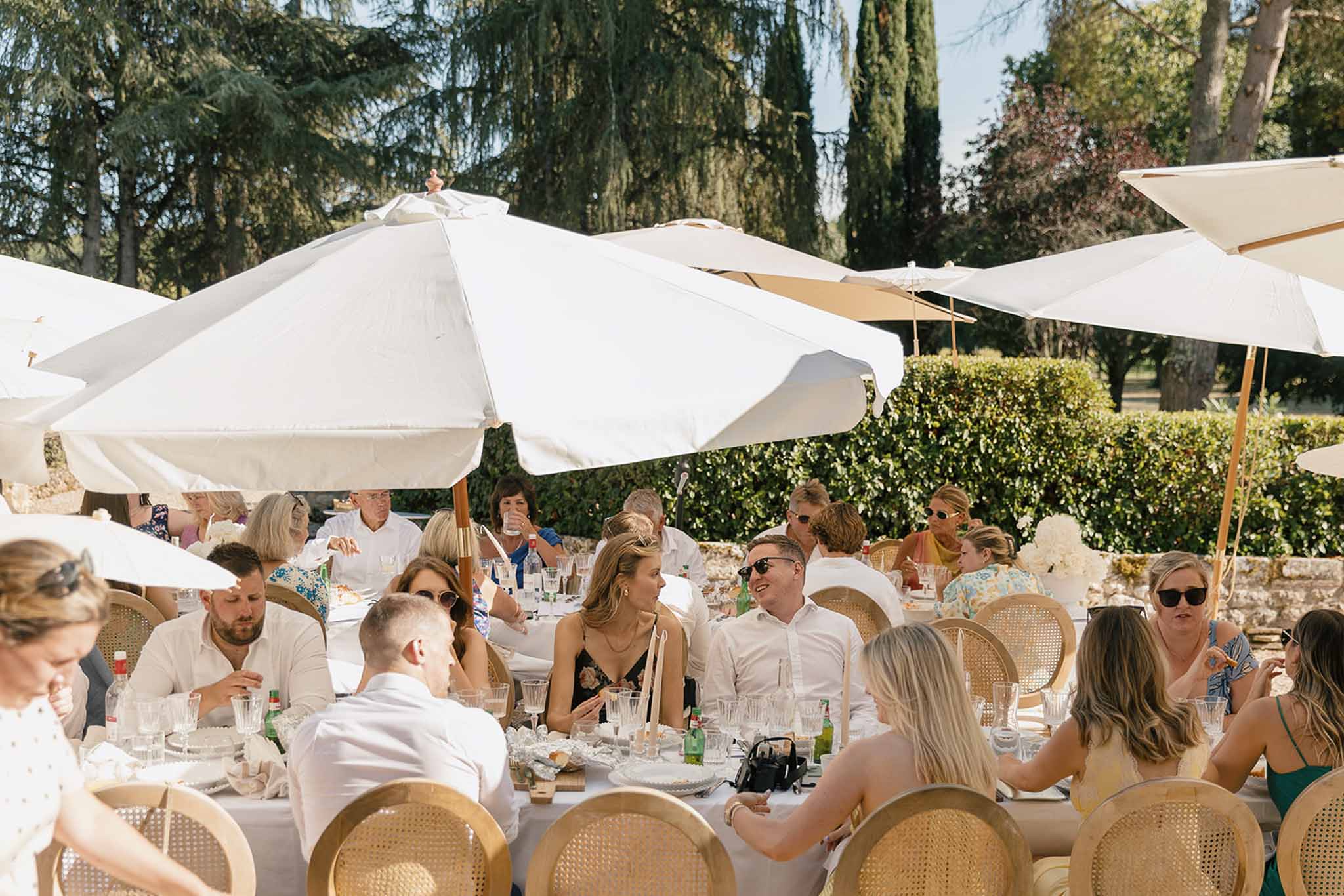 Guests seated at long white-linen tables with gold rattan chairs under white umbrellas in chateau garden