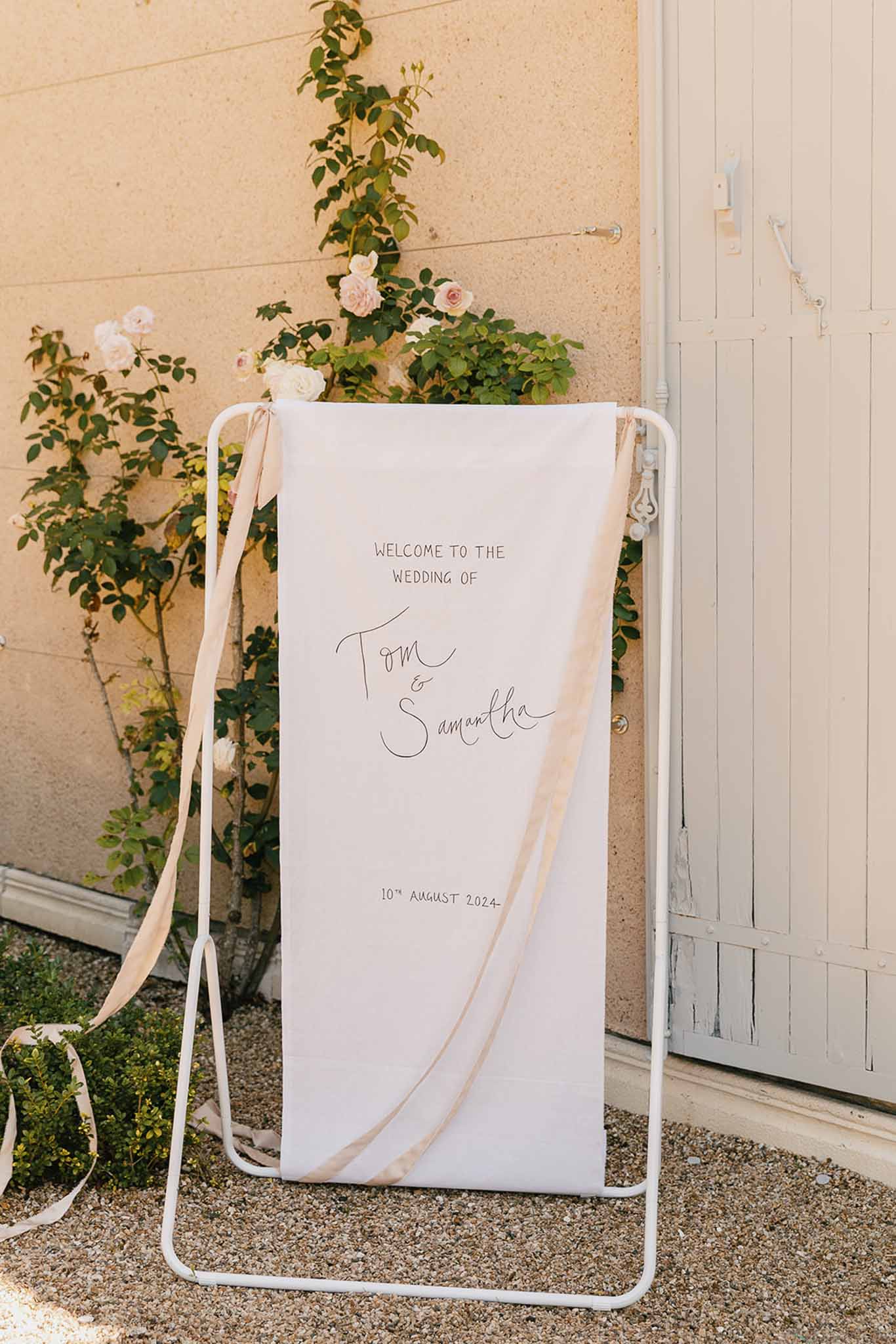 White fabric welcome sign with serif and script text on slim frame with trailing blush ribbons beside climbing roses