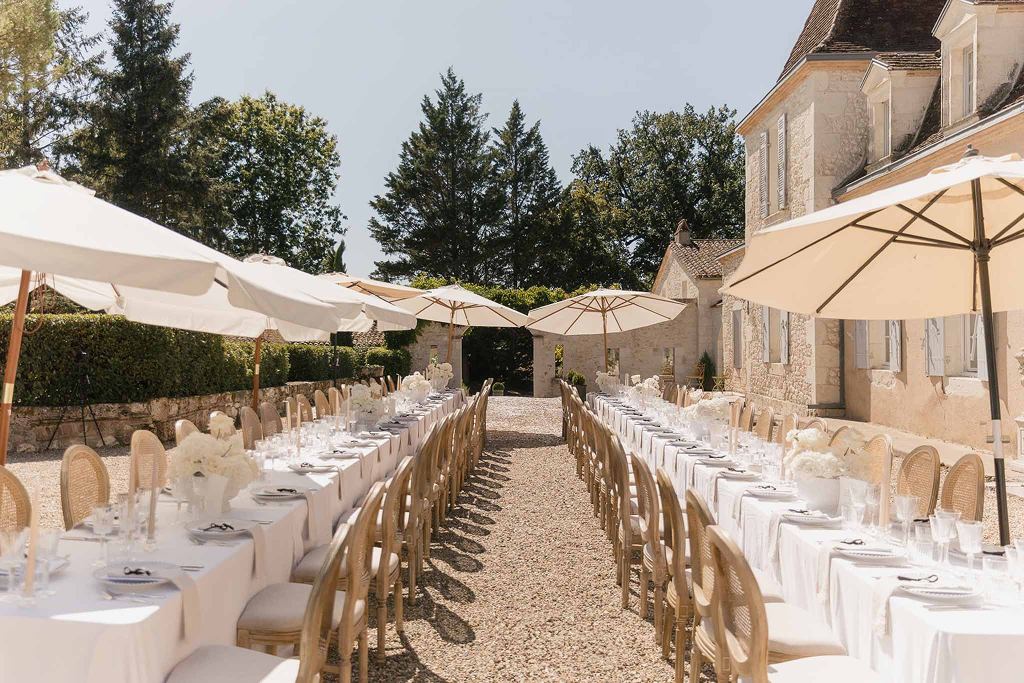 Outdoor wedding reception with long tables, white linens, and cream umbrellas in a French limestone manor courtyard