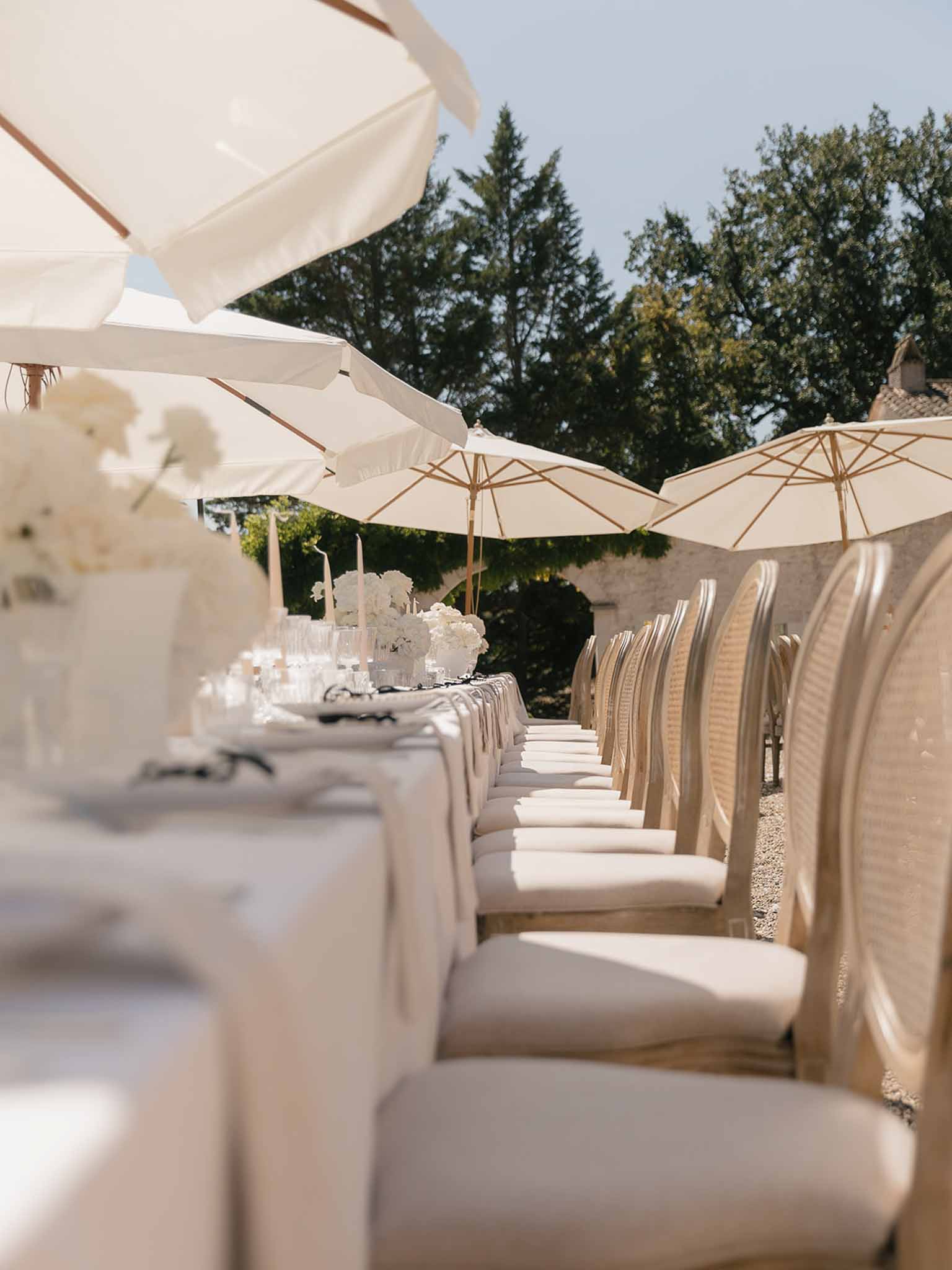 Long outdoor reception table with cream linens, white hydrangea centerpieces, taper candles, and market umbrellas in a sto...