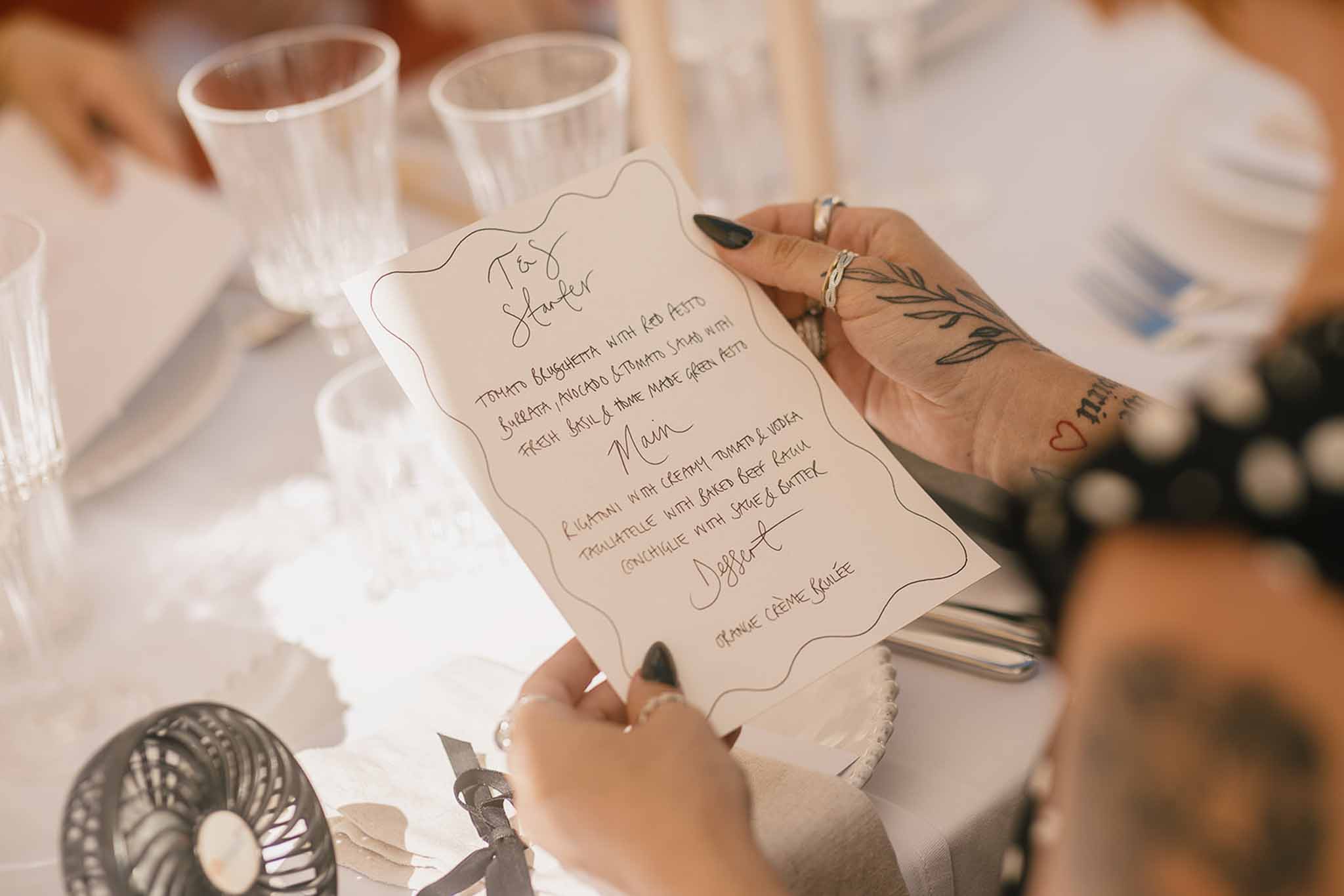 Guest holding handwritten calligraphy menu card with wavy border listing Italian courses at outdoor reception table