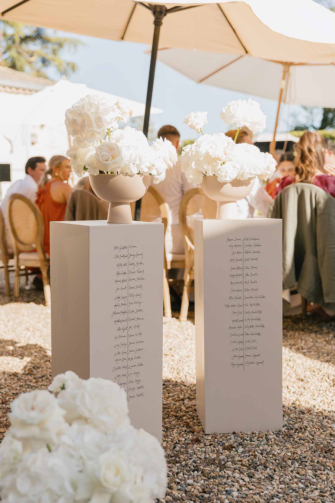 Two white plinths with calligraphy seating chart topped by sand ceramic bowls of white peonies on gravel terrace