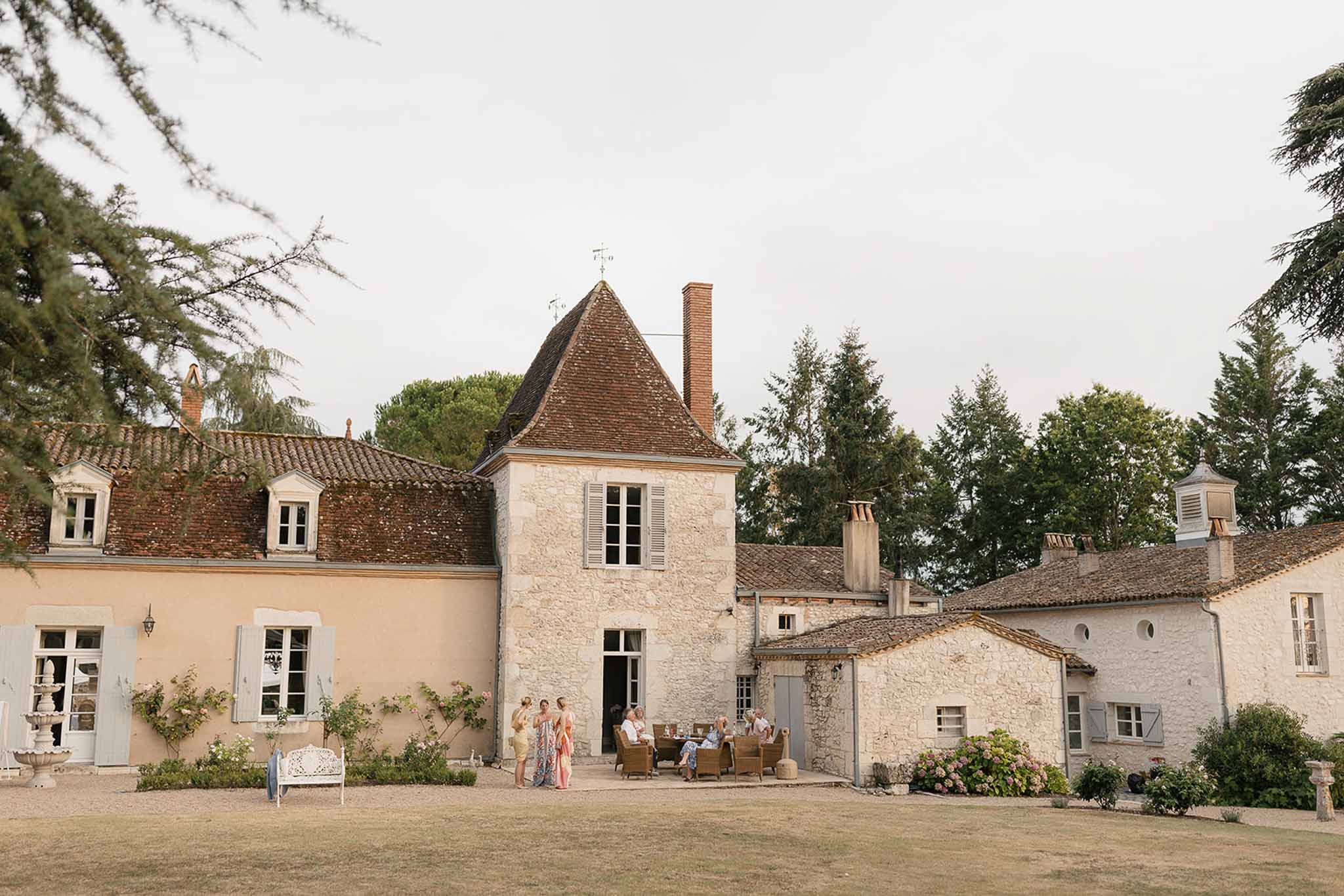 French country estate with turret and terracotta roof, guests on rattan furniture and stone fountain