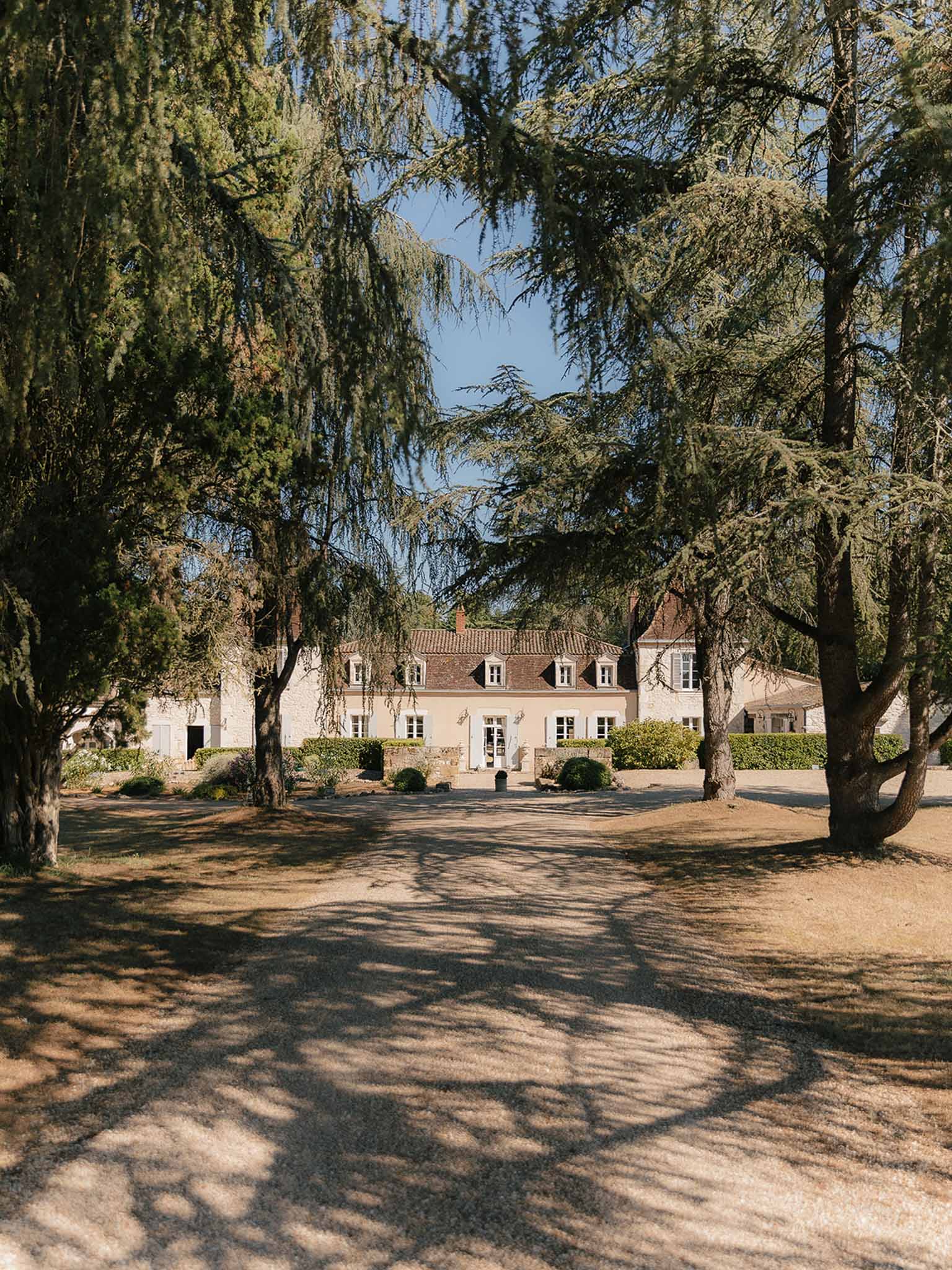 French country manor house with cream walls and dormer windows viewed from gravel driveway lined with cedar trees