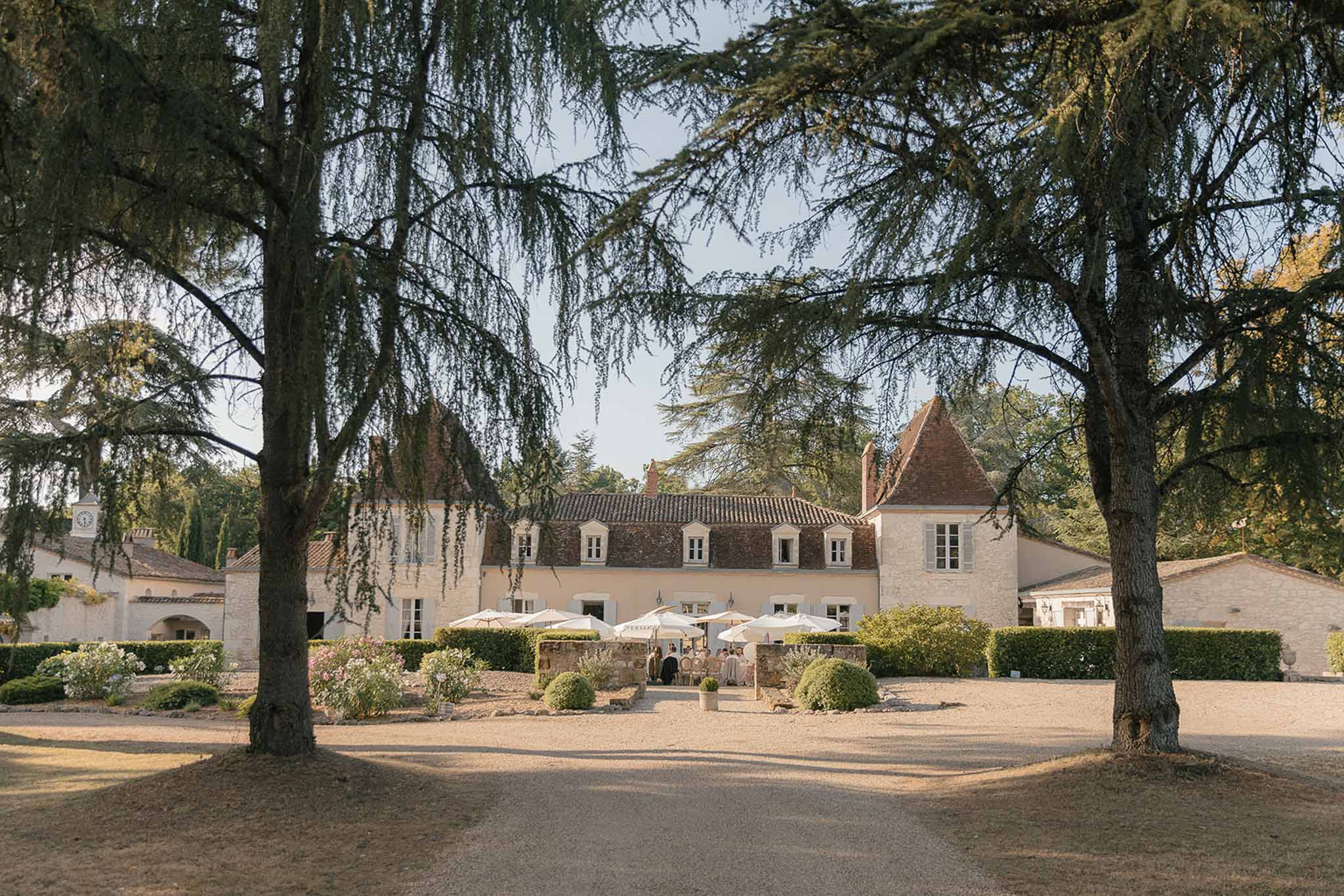 Turreted chateau with terracotta roof and white umbrellas on terrace framed by cedar trees