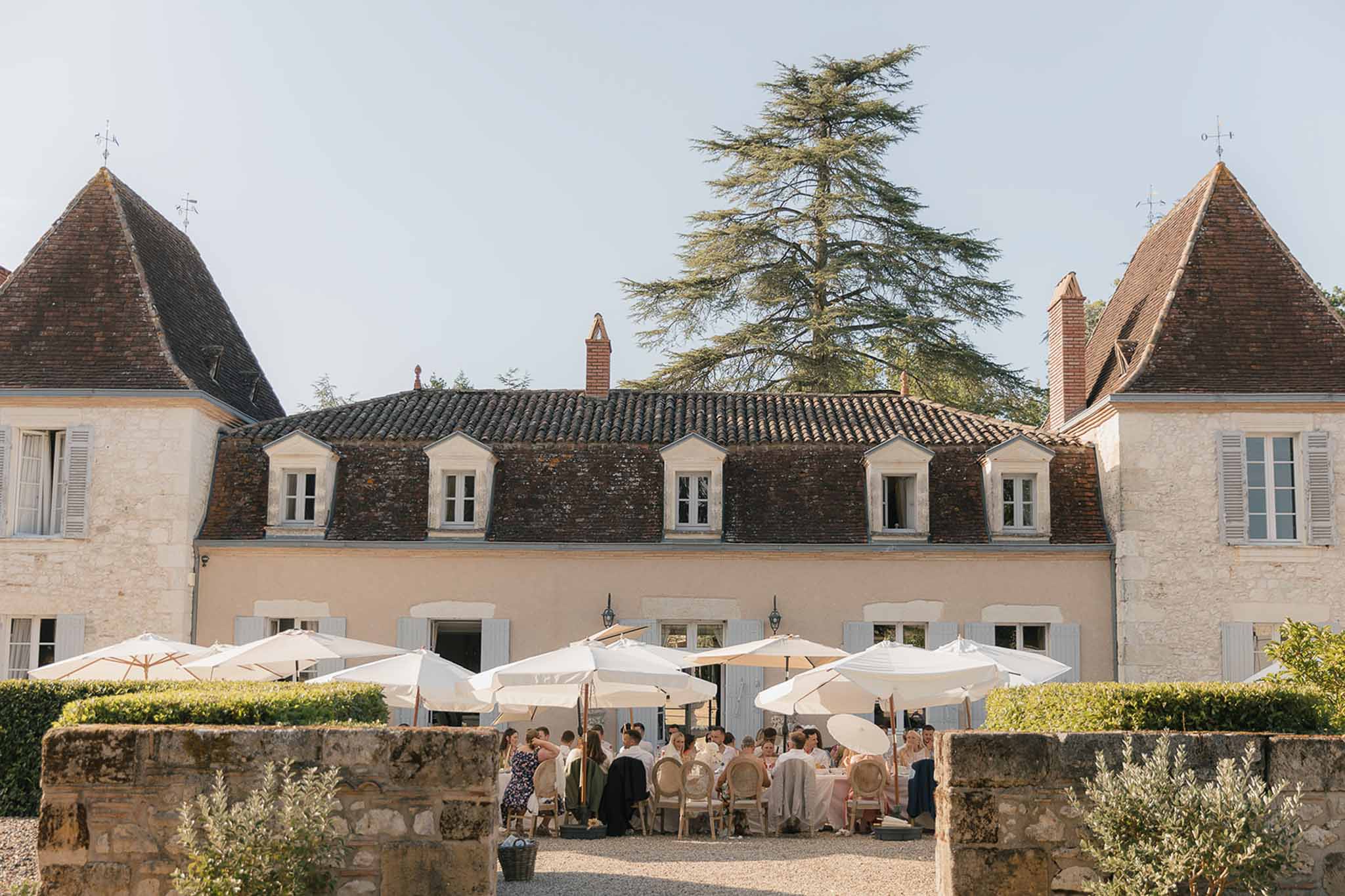 Guests at round white-draped tables under market umbrellas in chateau courtyard with pointed towers