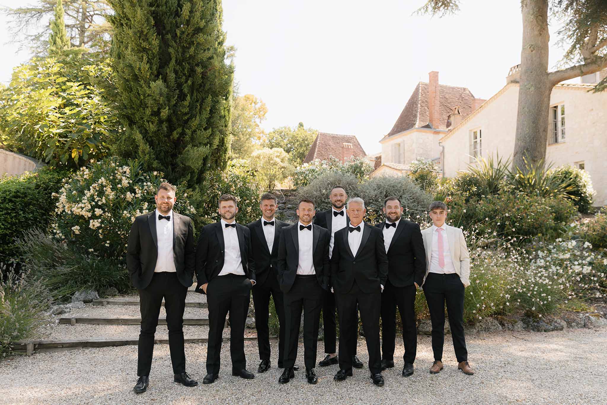 Eight men in matching black tuxedos posing on gravel path at French chateau estate