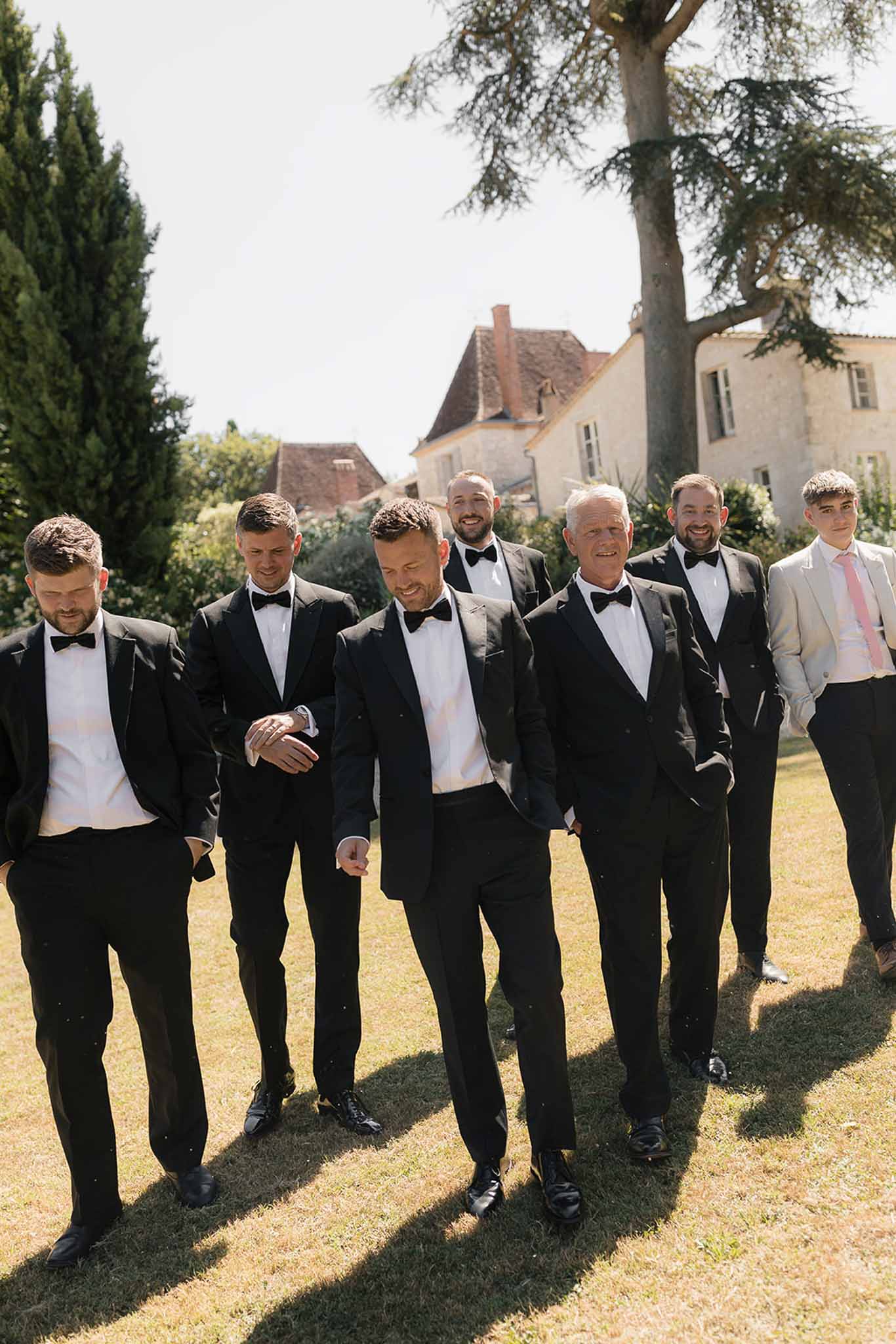 Groom and six groomsmen in black tuxedos walking across chateau lawn in bright sunlight