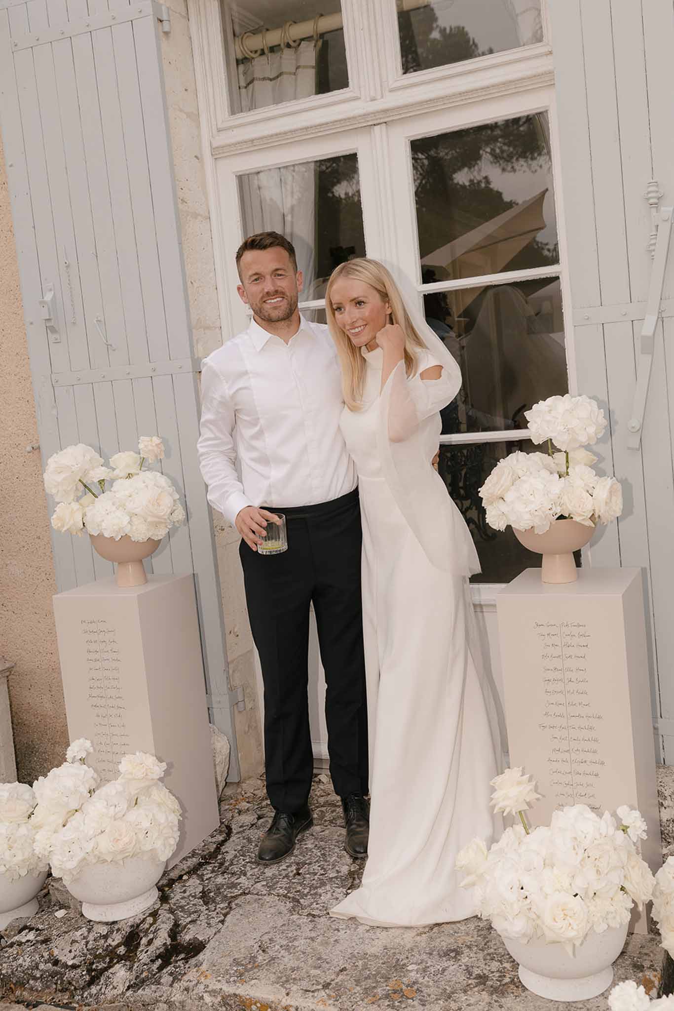 Bride in minimalist ivory gown and groom with suspenders between white peony arrangements at chateau entrance