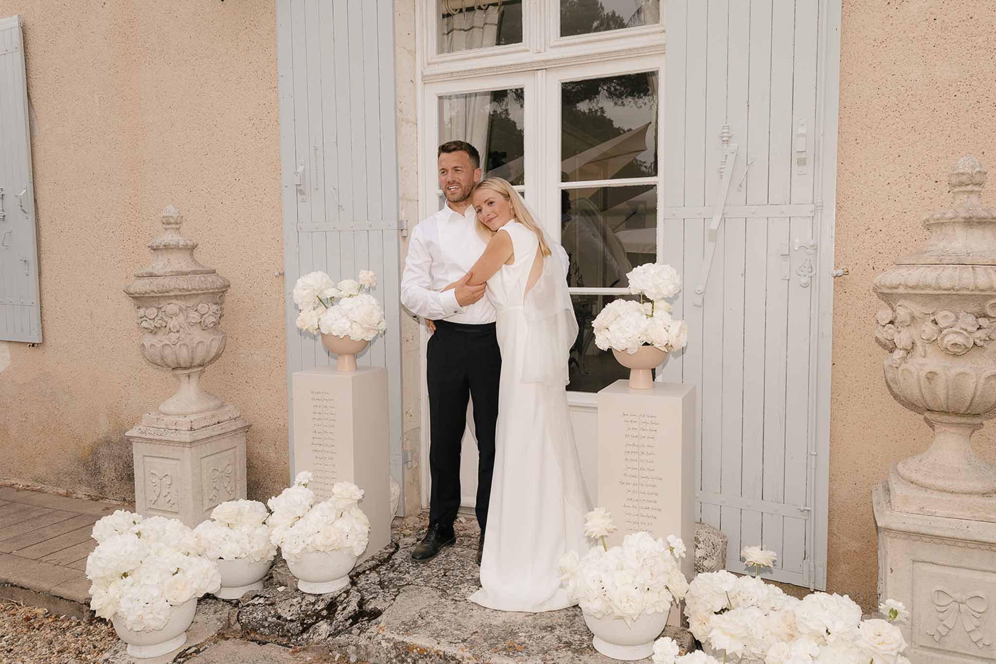 Couple embracing before all-white hydrangea and peony entrance installation with seating chart pedestals