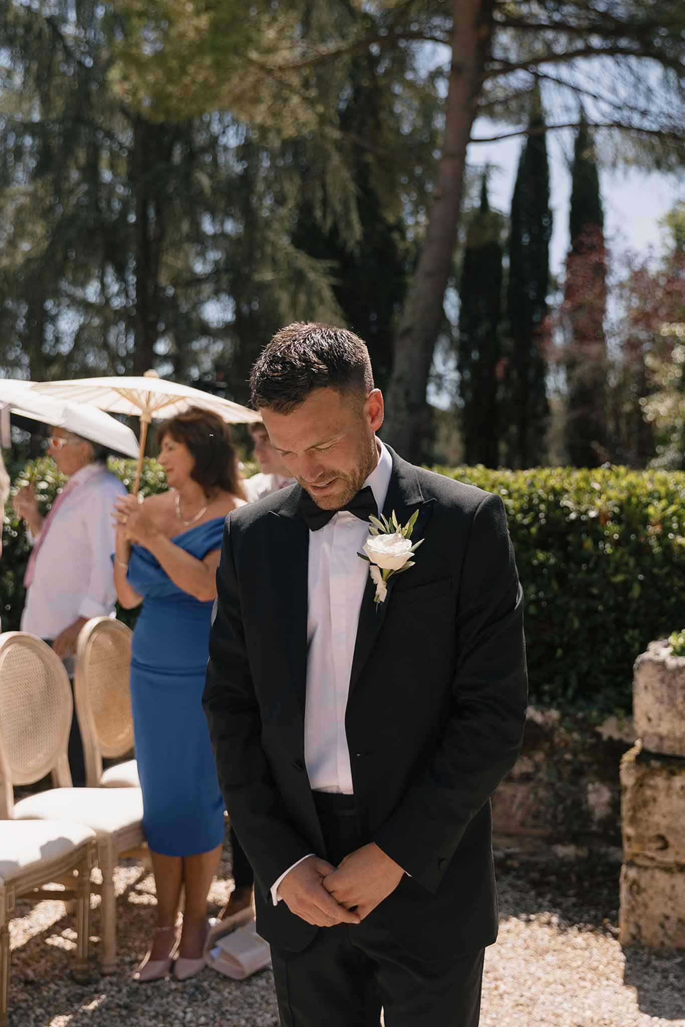 Emotional groom in black tuxedo with white rose boutonniere standing at outdoor garden ceremony altar