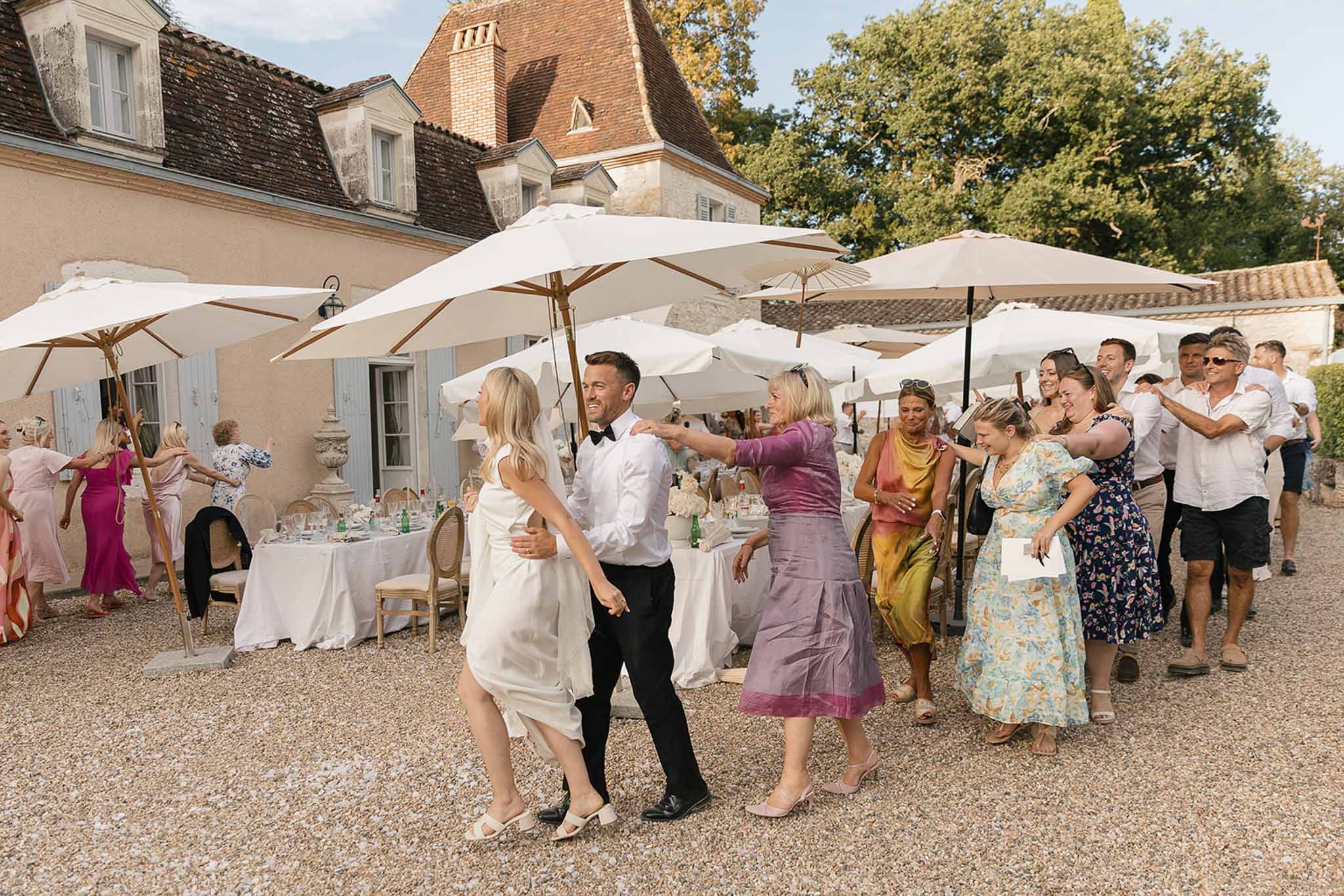 Couple dancing on gravel courtyard as guests mingle under white umbrellas at pink-walled manor