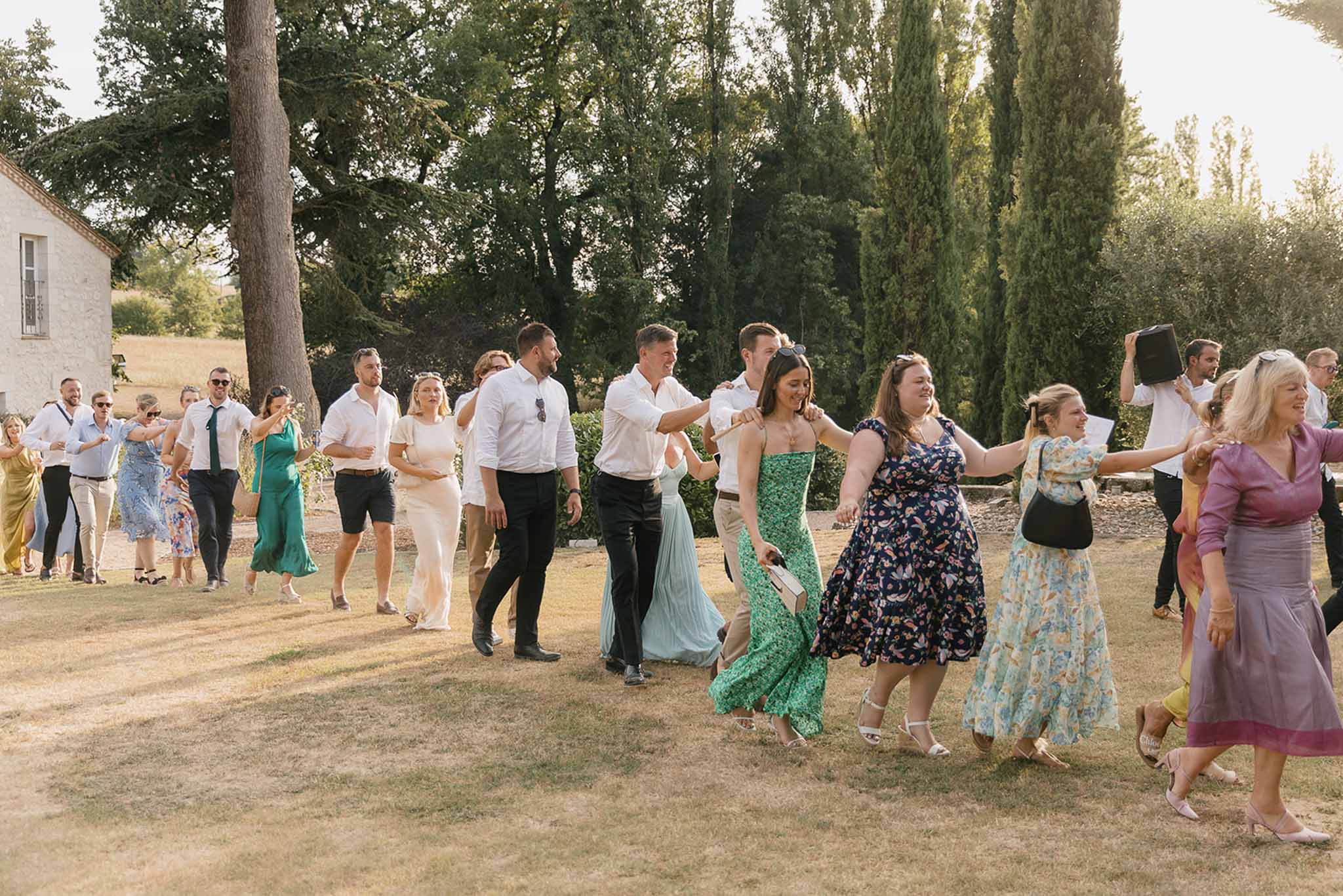Twenty-five guests in conga line on golden-hour estate lawn with cypress trees in background