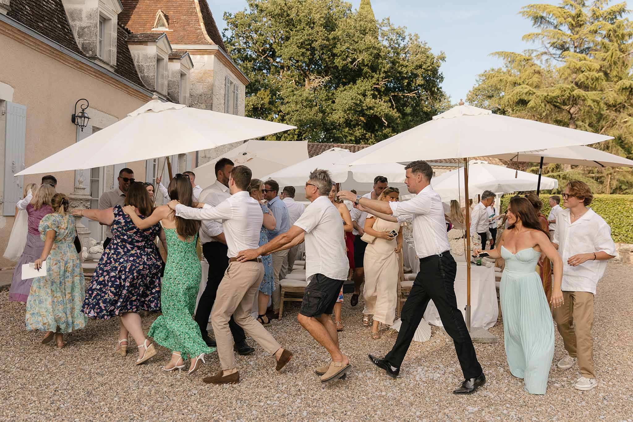 20 guests dancing in conga line on gravel terrace under white market umbrellas beside stone manor