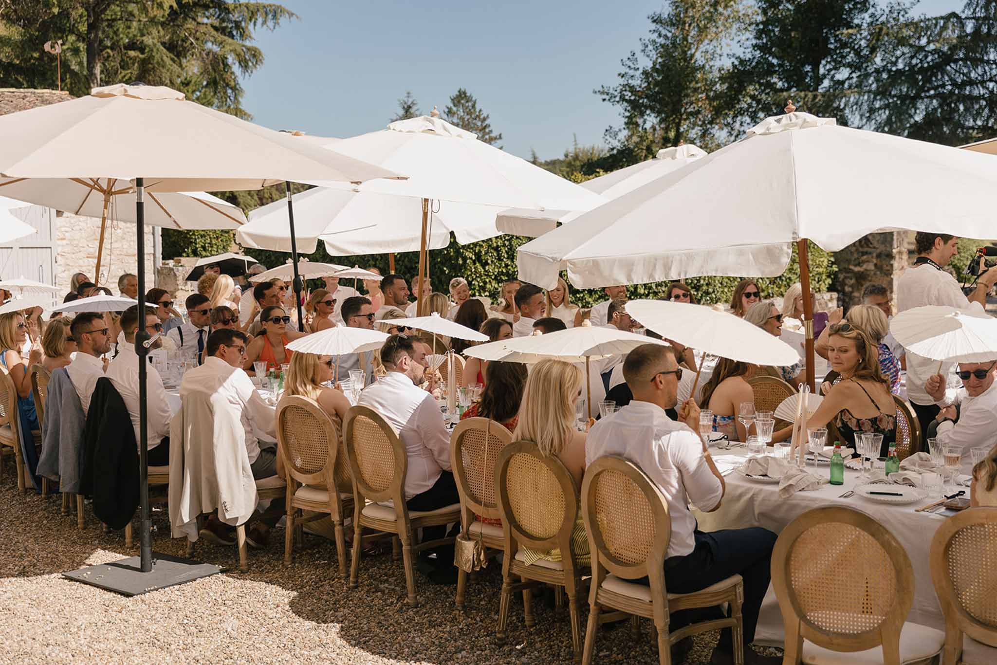 Forty guests at long white-draped tables under market umbrellas with rattan Louis chairs in sunny courtyard