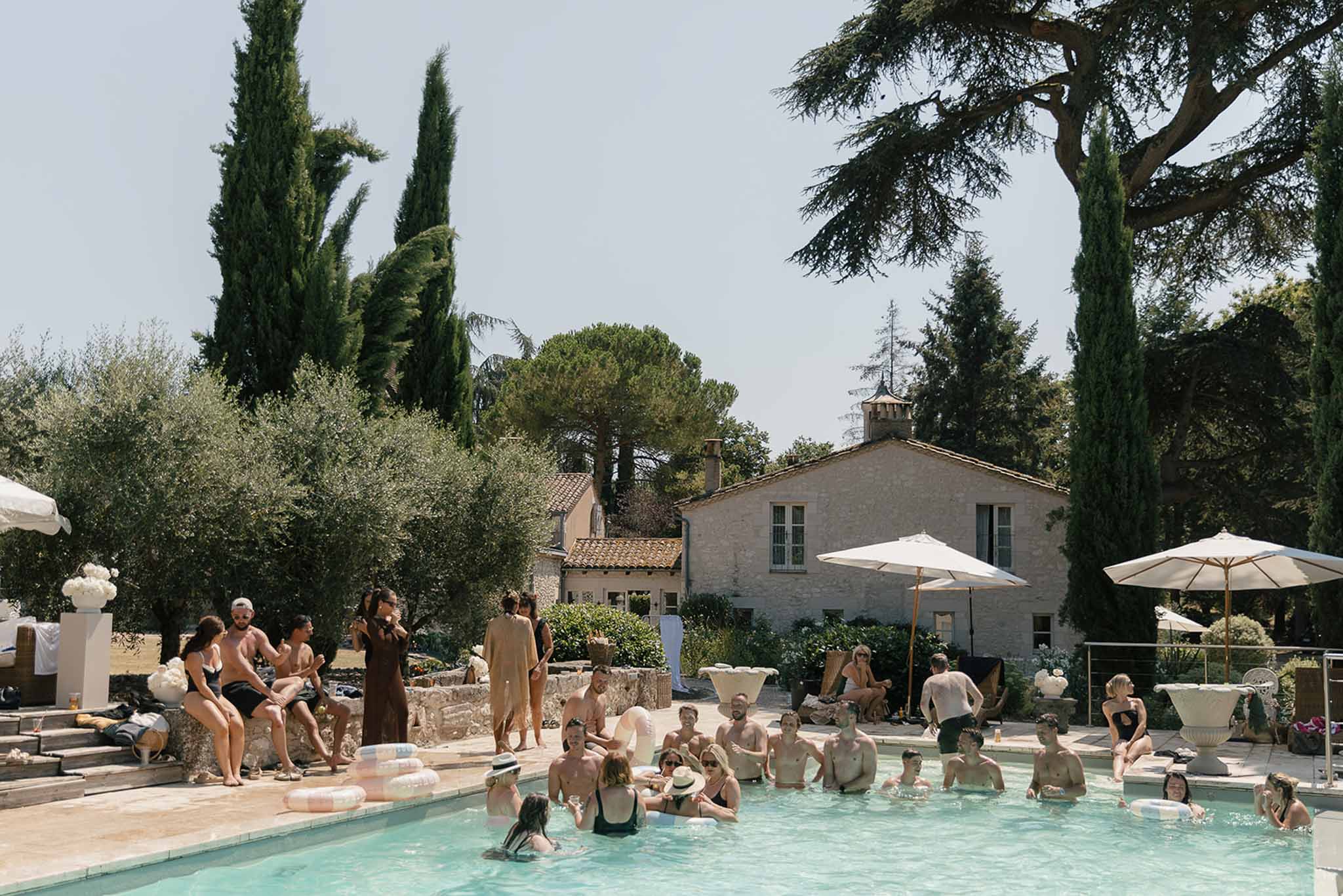 Wedding guests socializing in and around pool at stone bastide property with cream parasols and white florals