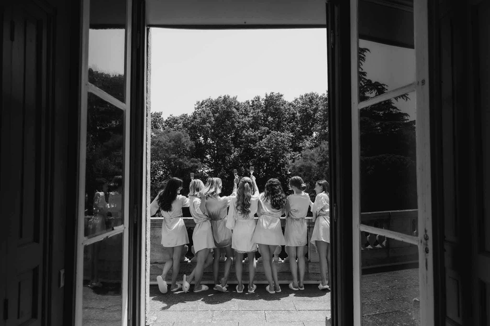 Seven women in matching robes raise champagne on stone balcony framed through open French doors in B&W
