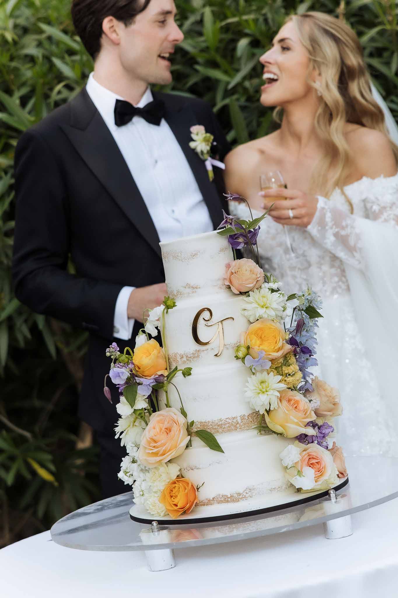 Four-tier semi-naked wedding cake with gold bands and cascading fresh flowers as couple laughs in soft focus behind