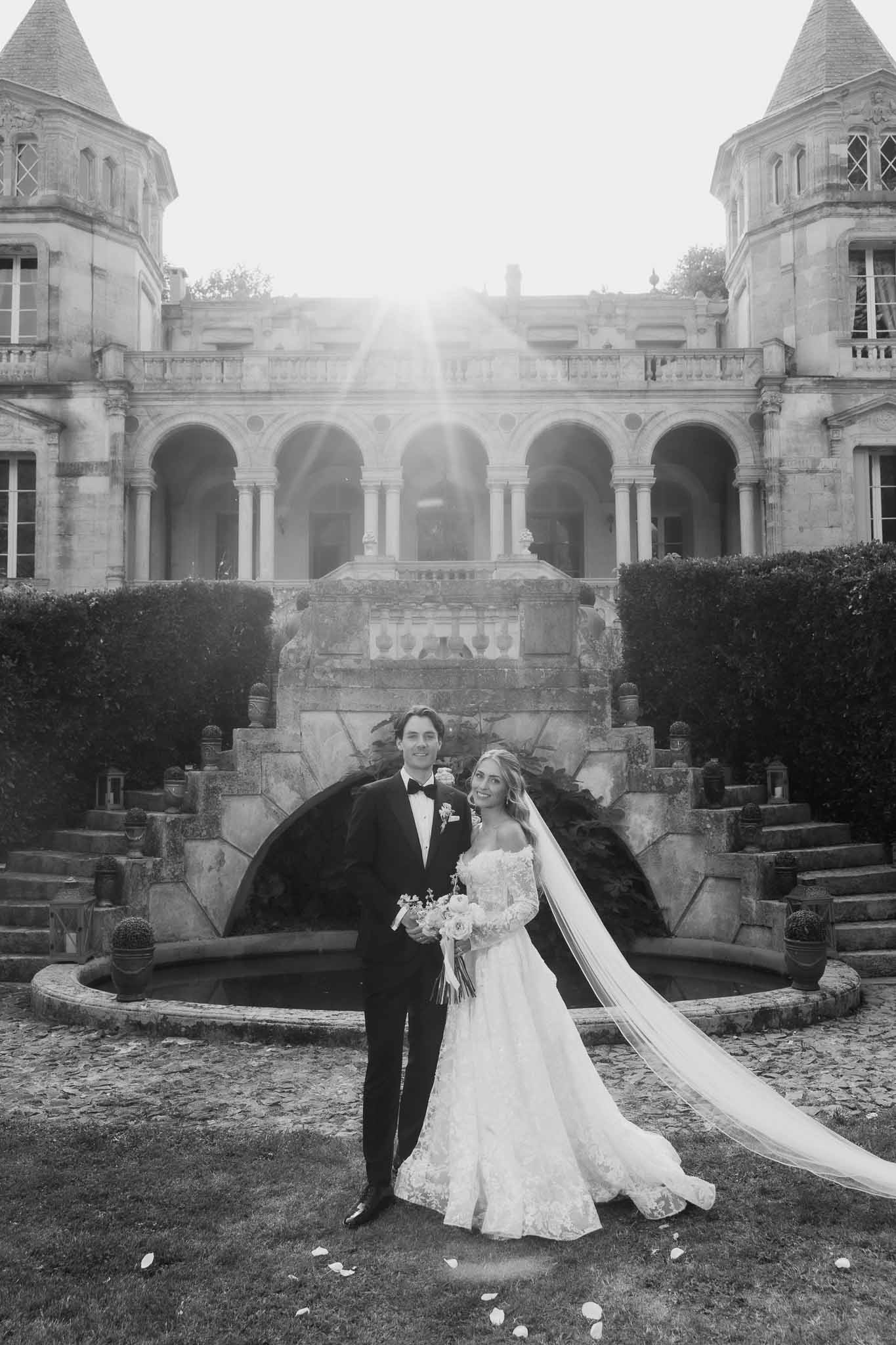 Black and white portrait of bride and groom in front of twin-turret chateau with cathedral veil and sun flare