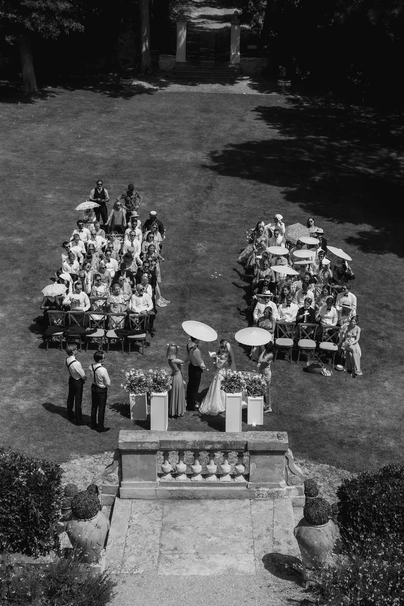 Black-and-white aerial view of outdoor wedding ceremony on chateau garden lawn with guests holding parasols