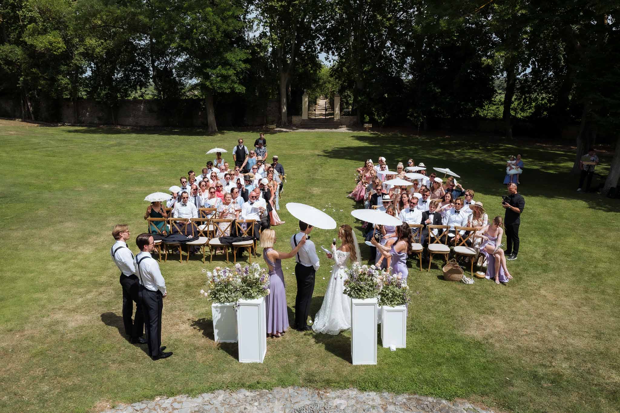 Elevated view of lawn ceremony with wildflower pedestals, cross-back chairs, and guests holding parasols