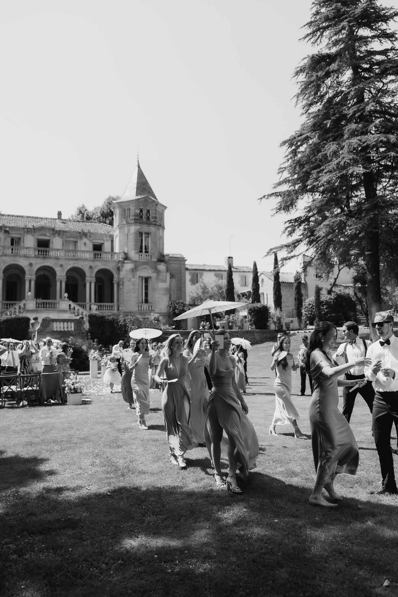 Black-and-white wide shot of bridal party and guests walking across chateau lawn after ceremony with parasols