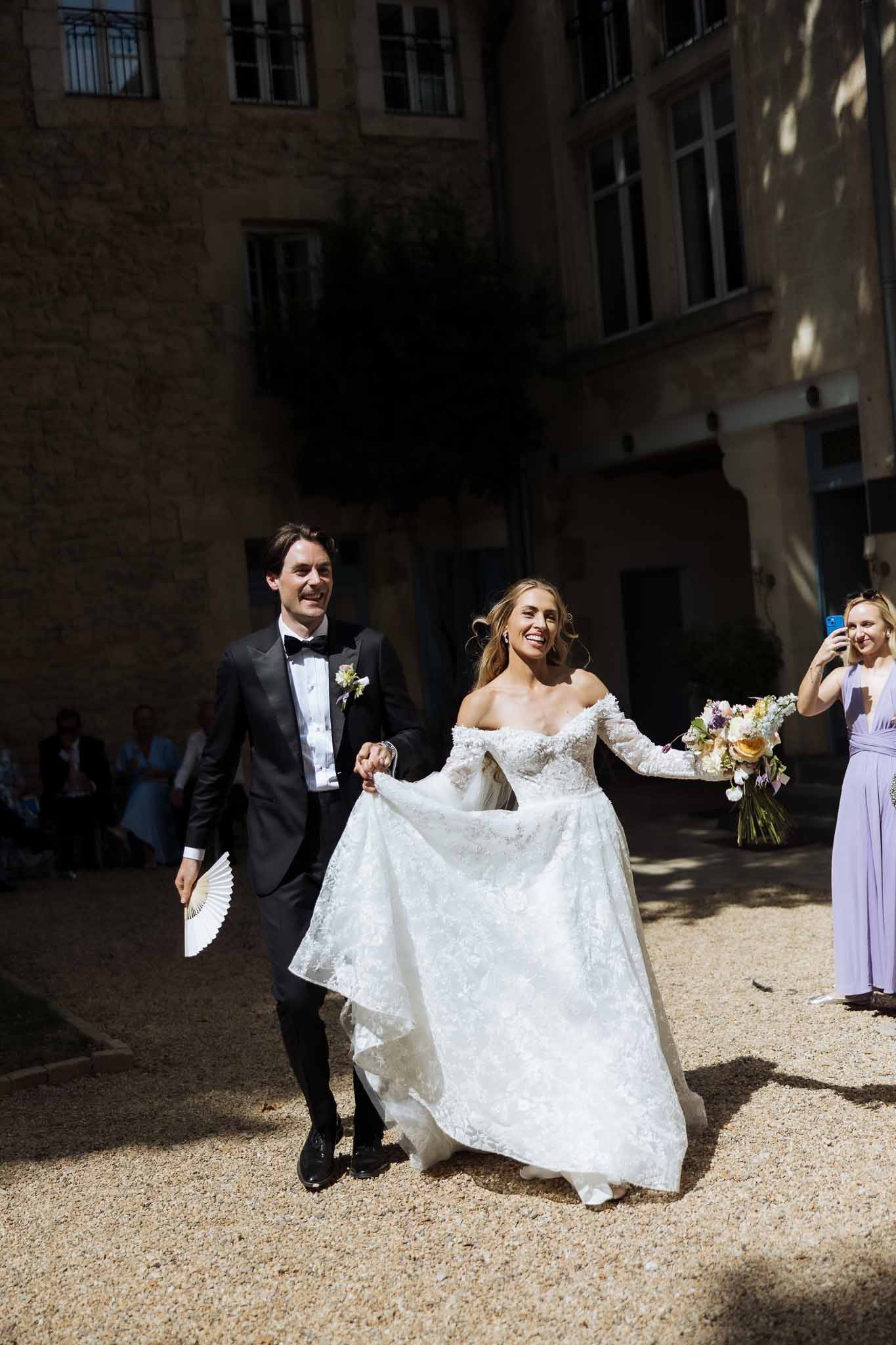 Bride in off-shoulder lace ballgown with peach and lavender bouquet walking hand-in-hand with groom on gravel courtyard