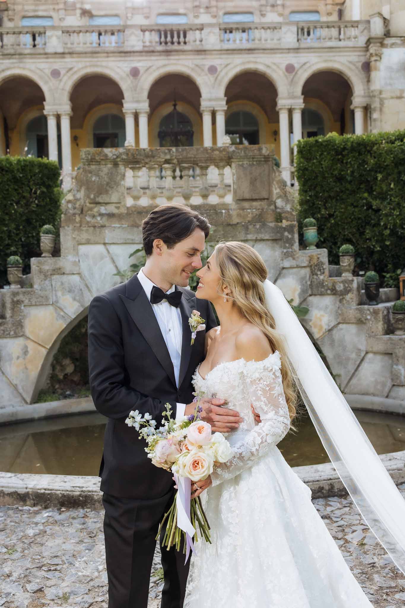 Bride in white lace gown and cathedral veil with groom in black tuxedo facing each other in chateau courtyard