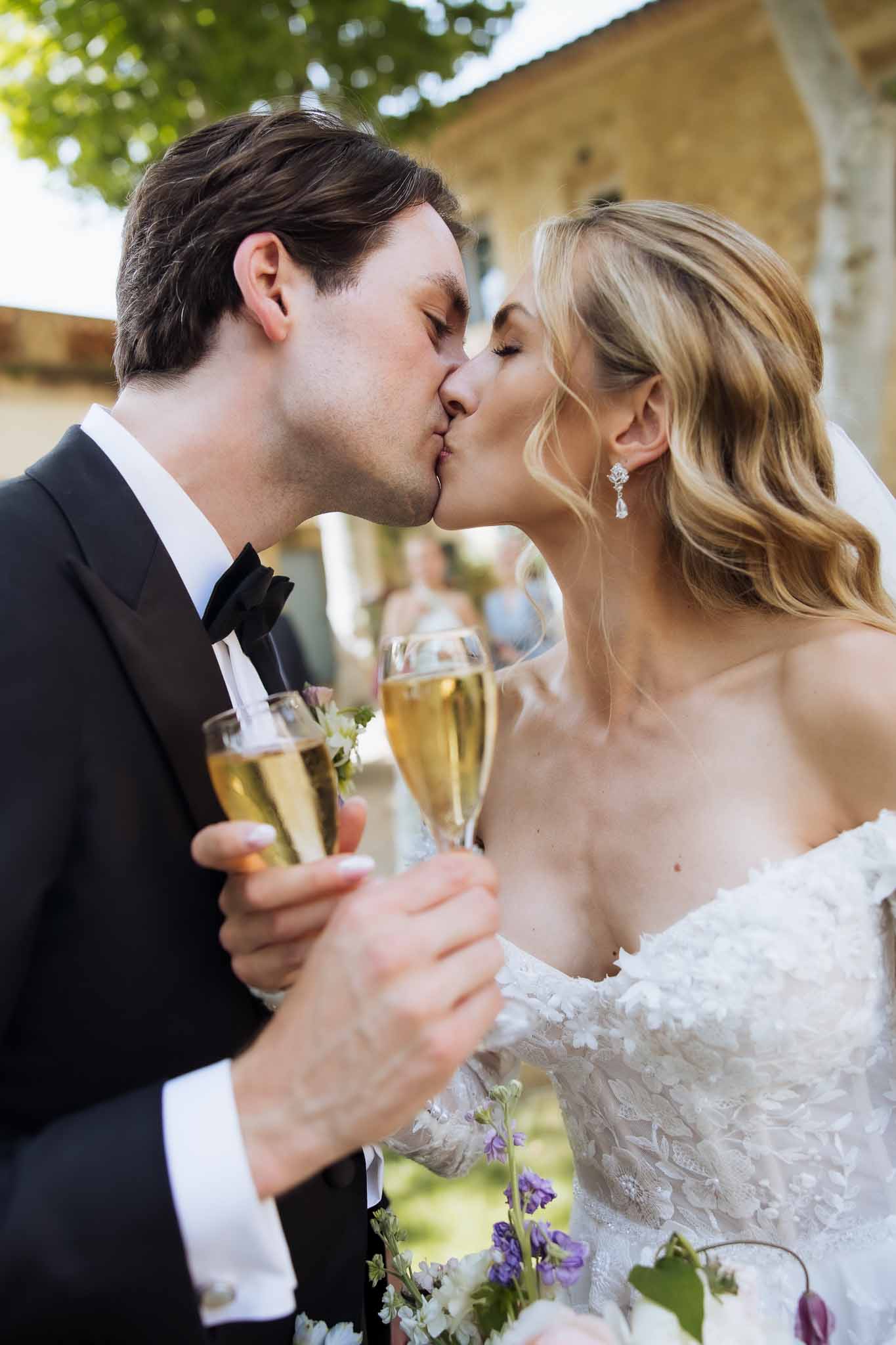 Bride in lace applique gown with purple delphinium bouquet kissing groom while both hold champagne flutes