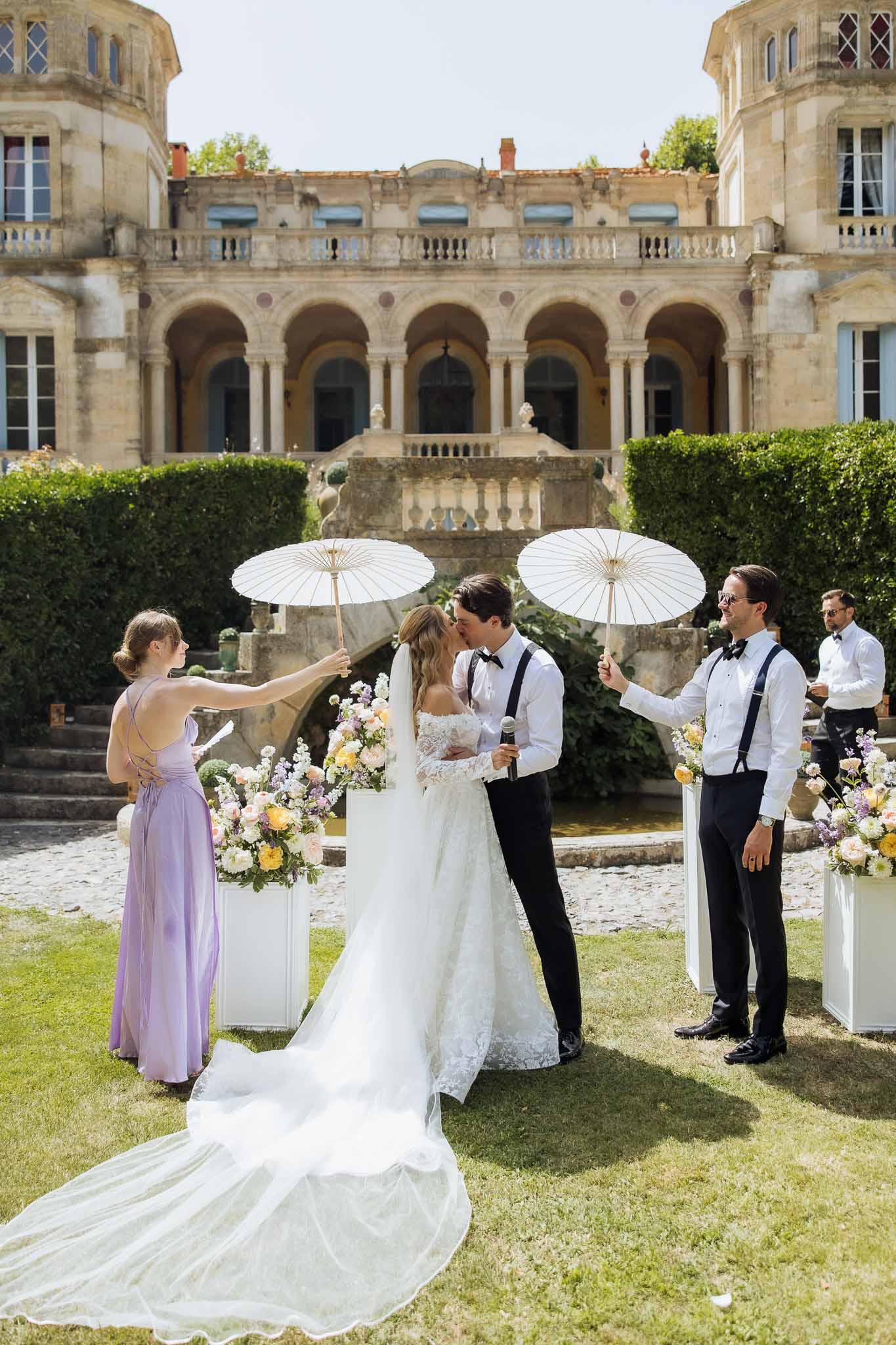 Bride and groom share first kiss under parasols at chateau ceremony with pastel floral arrangements