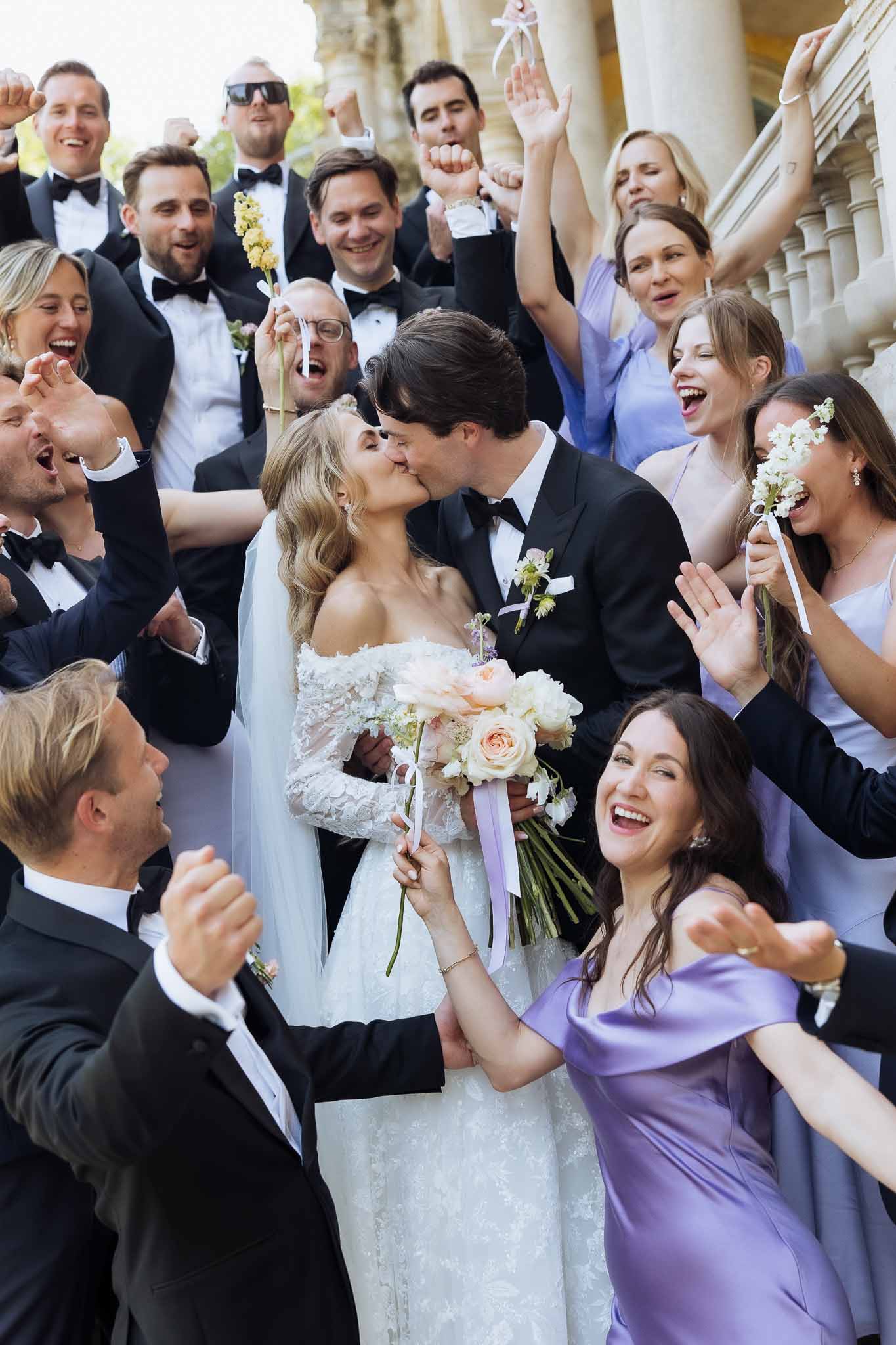 Couple kissing amid cheering bridal party with lavender bridesmaids and black-tie groomsmen on classical stone steps