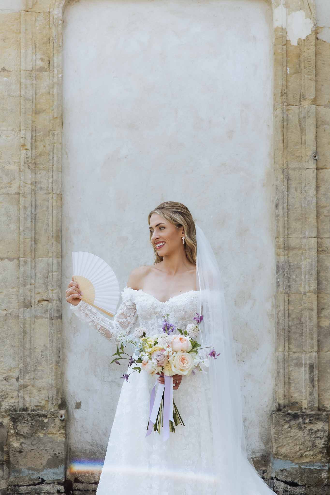 Bride in off-shoulder lace gown holding peach and mauve bouquet with paper fan before stone arched doorway